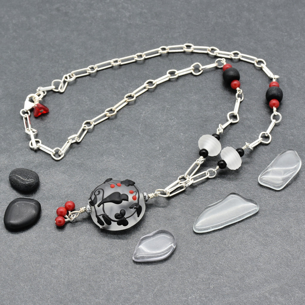 A silver-toned chain necklace with a round black and gray patterned pendant, accented by small red beads displayed on a dark gray textured background - made with love in Colorado by Ruby Acorn.