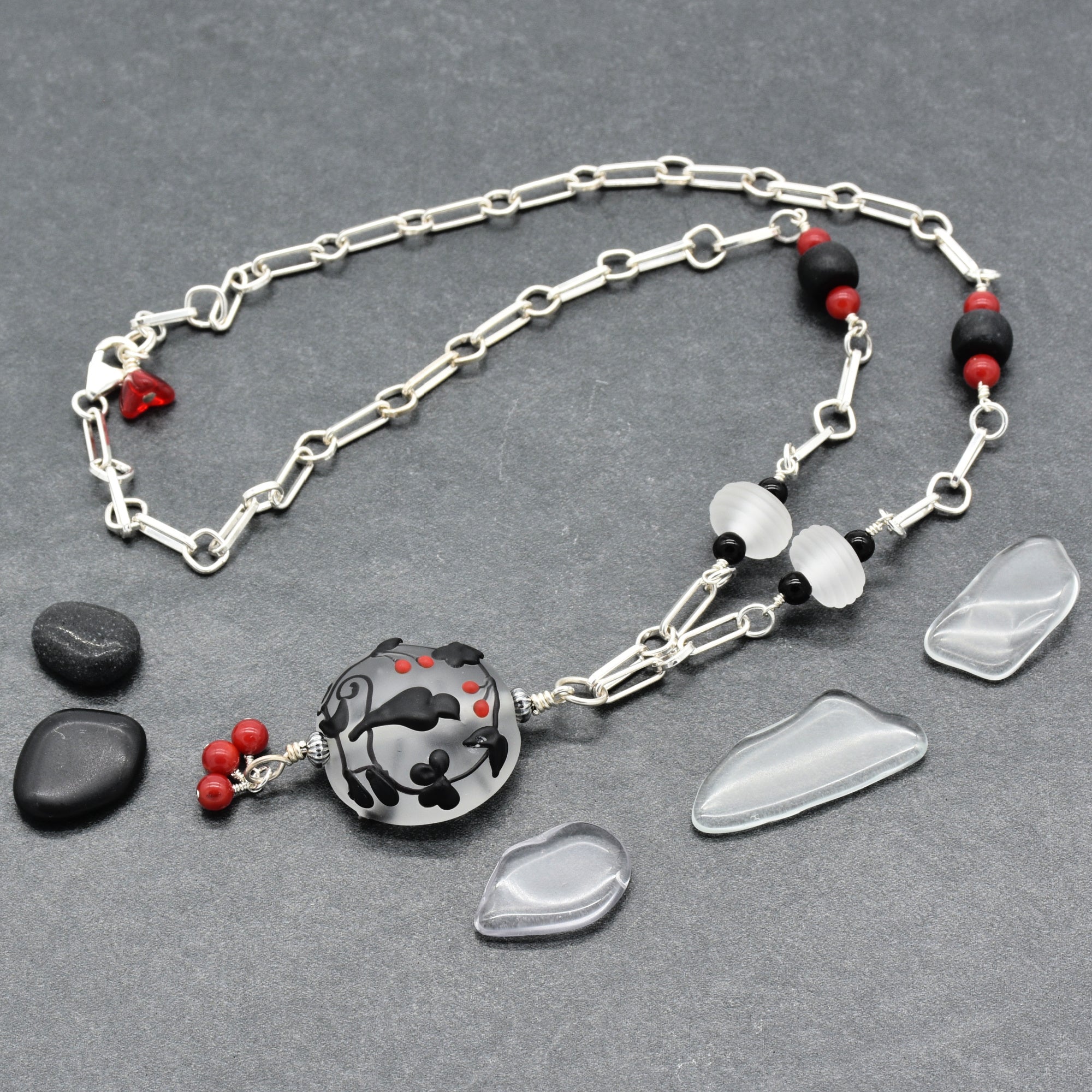 A silver-toned chain necklace with a round black and gray patterned pendant, accented by small red beads displayed on a dark gray textured background - made with love in Colorado by Ruby Acorn.