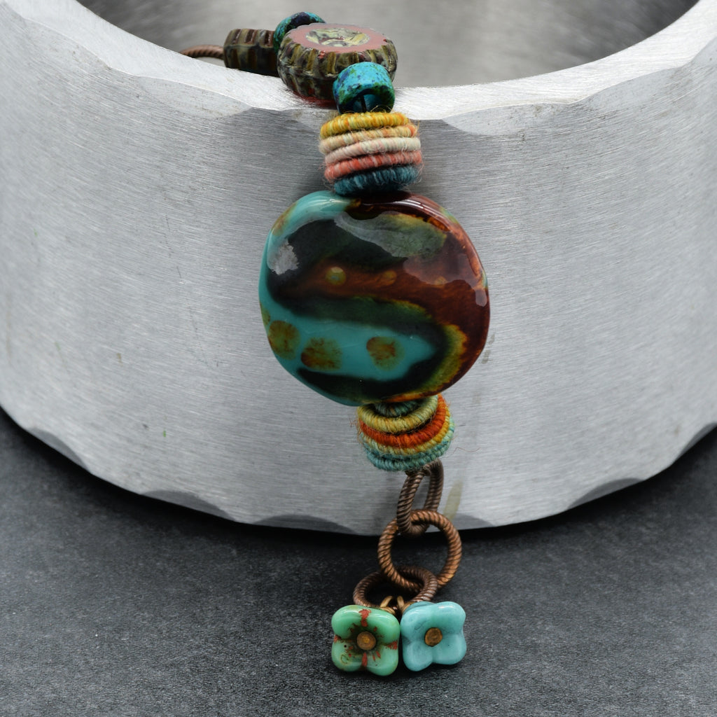 This image shows a bracelet. The jewelry piece is small in size and has an opening clasp, which are characteristic features of a bracelet. It has a round multicolored central bead, smaller colorful beads, and two small flower charms at the end, all strung together on a cord.