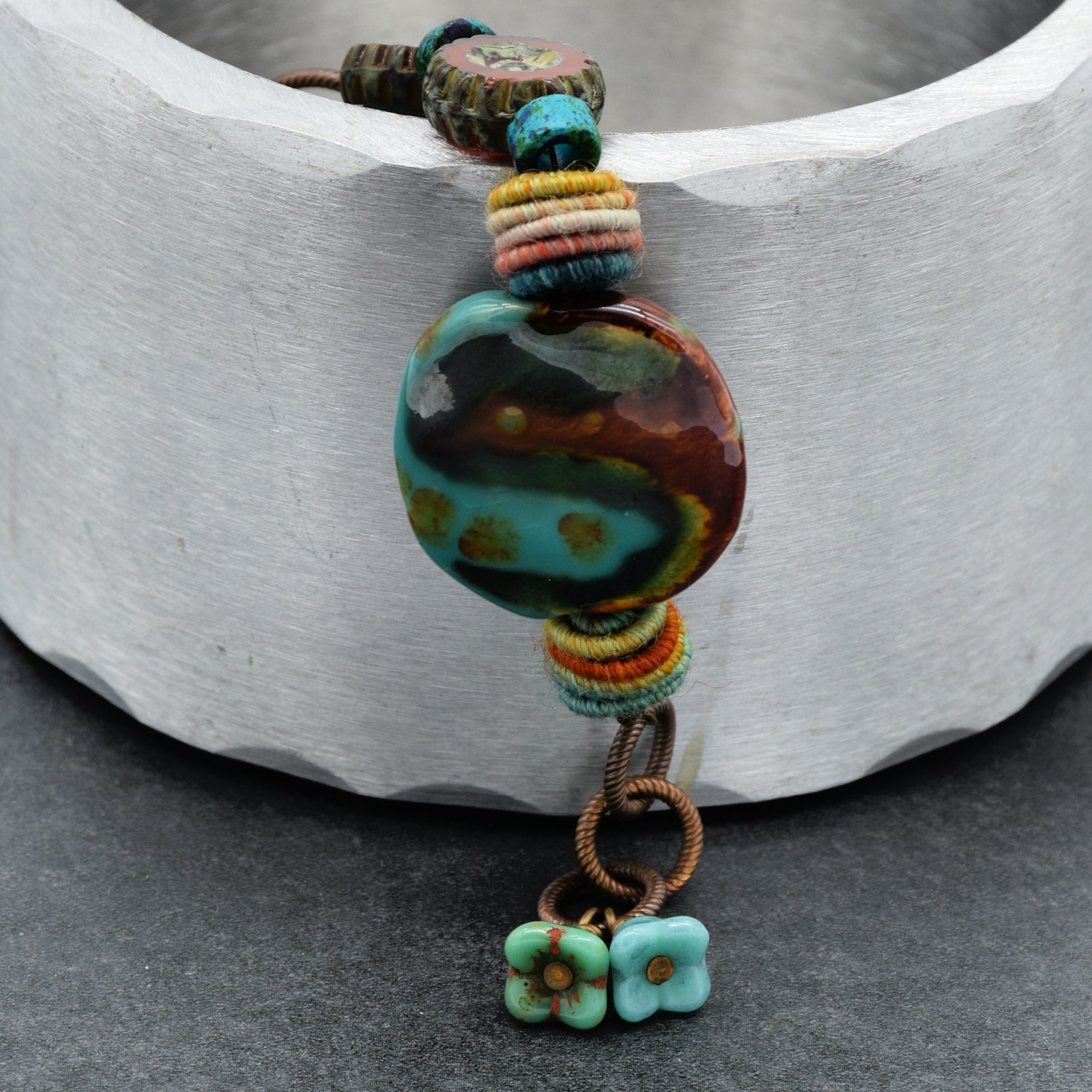 This image shows a bracelet. The jewelry piece is small in size and has an opening clasp, which are characteristic features of a bracelet. It has a round multicolored central bead, smaller colorful beads, and two small flower charms at the end, all strung together on a cord.