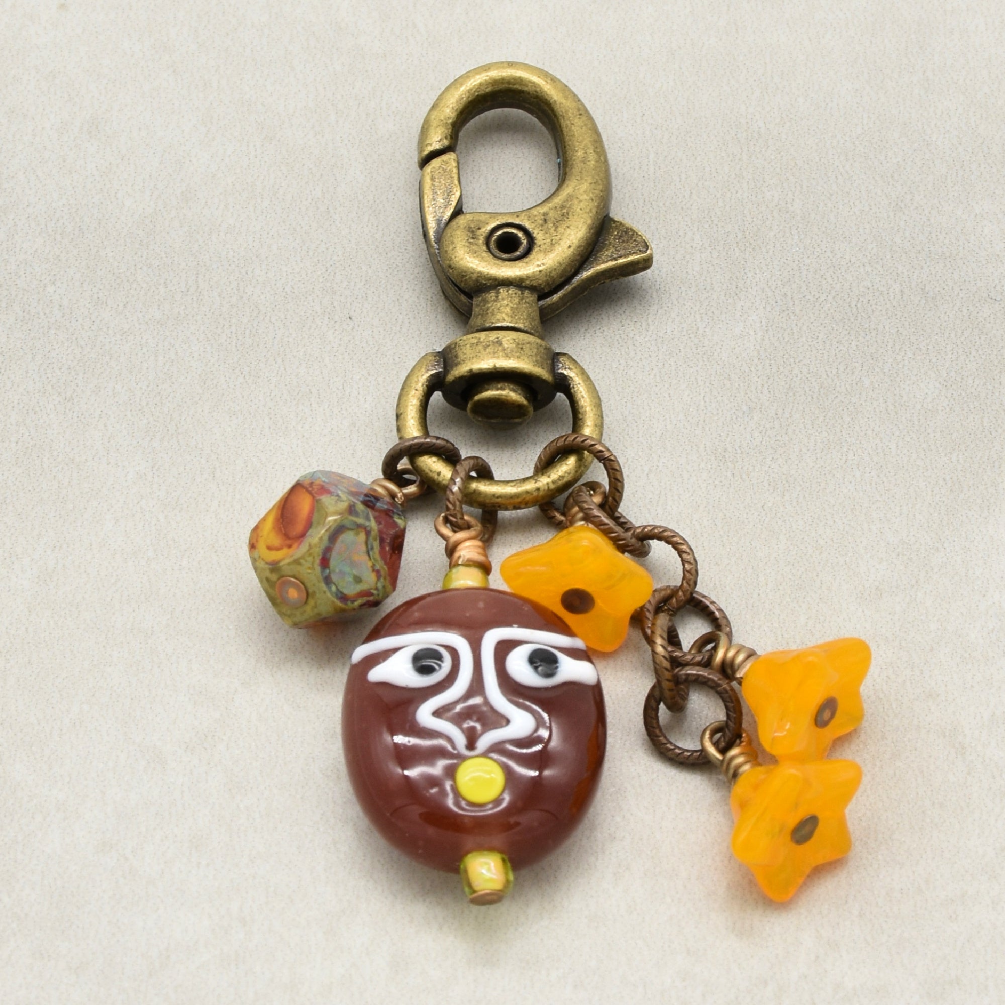 An antique bronze-finished lobster clasp keychain featuring a brown glazed face bead with white painted details, a multi-colored patterned bead, and three small yellow bellflower-shaped beads on a light gray background - made with love by Ruby Acorn.