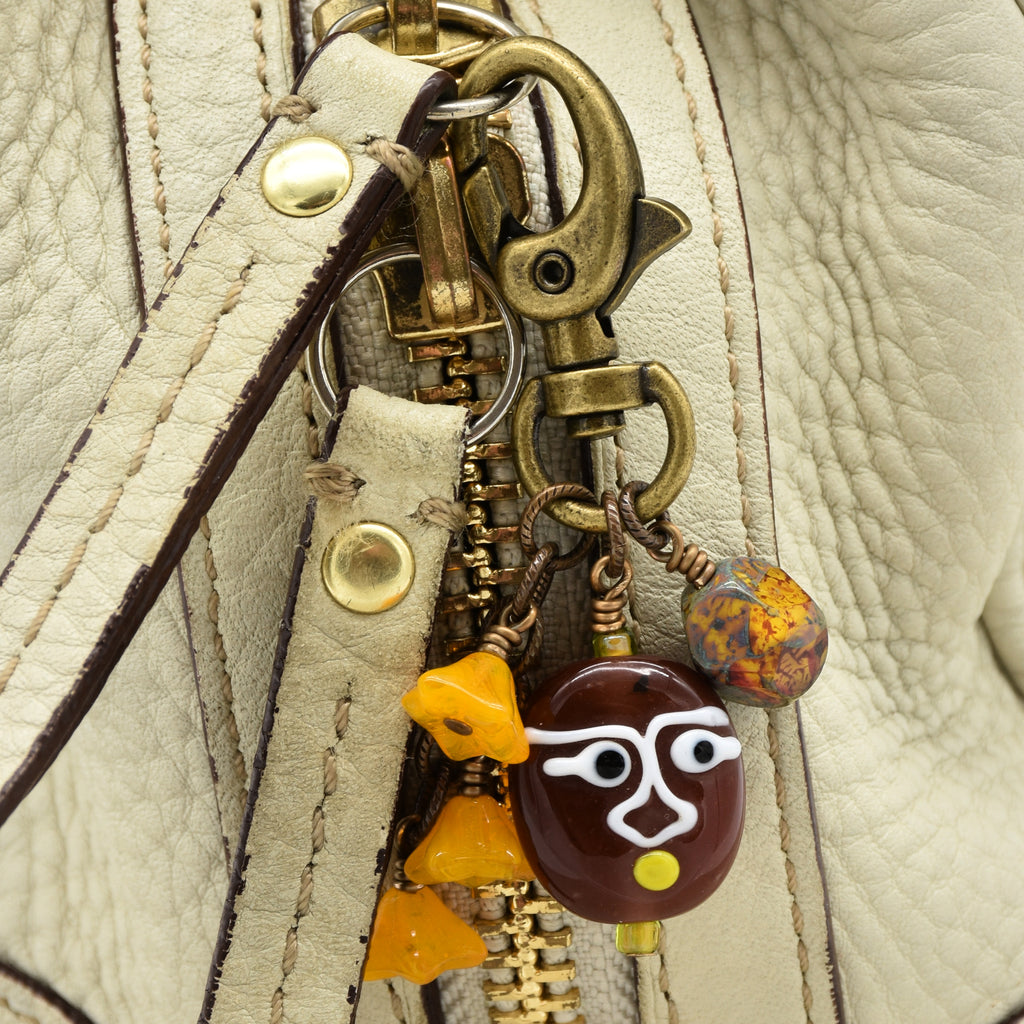 Close-up of a cream-colored textured leather handbag featuring a brass-tone zipper pull adorned with decorative glass charms, including a brown face pendant and orange-yellow beads - made with love in Colorado by Ruby Acorn.