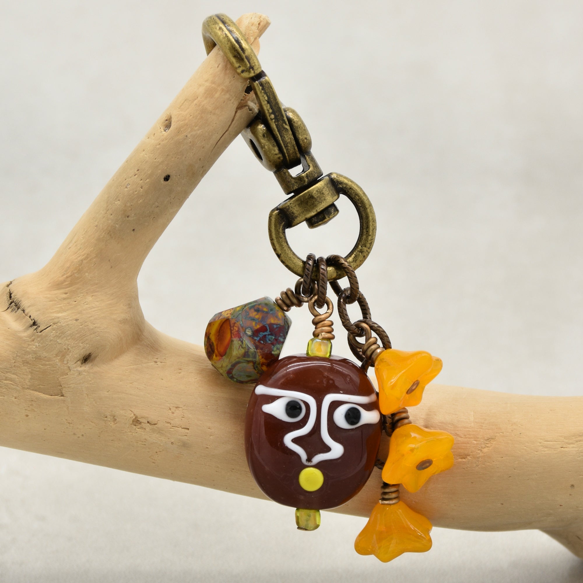 A close-up of a jewelry keychain or bag charm draped over a light wood branch. The charm features an antique bronze metal clasp and chain, a dark brown round bead with a white and yellow painted face, a small multicolor bead, and three small yellow flower-shaped beads - made with love by Ruby Acorn.
