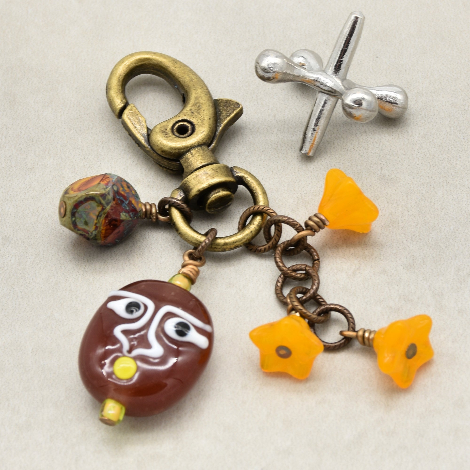 A handmade keychain or bag charm featuring a brass-colored lobster clasp, a brown glossy beaded mask charm, three bright yellow flower charms and a marbled bead displayed on a light beige background - made in Colorado with love by Ruby Acorn.