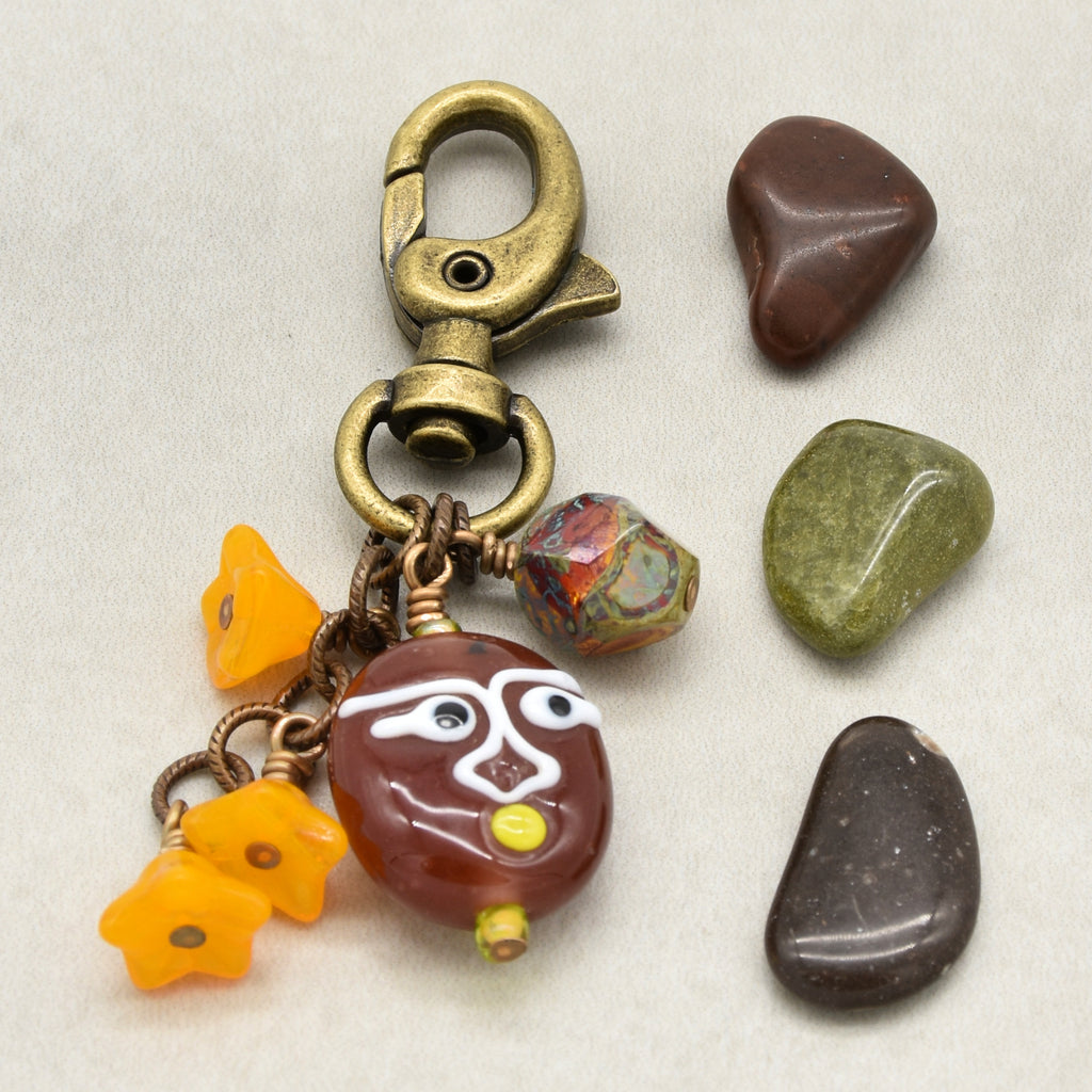A handmade beaded keychain or bag charm with an antique brass lobster clasp, featuring a brown glass bead with a white face, yellow accents, and three small yellow flower beads, displayed next to four smooth river stones on a light gray background - made with love in Colorado by Ruby Acorn.