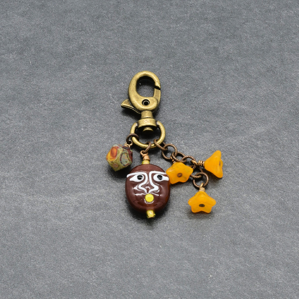 A small jewelry charm or keychain clasp with a lobster clasp, chain links, and three dangling charms: a brown oval bead with white painted facial features, three yellow flower-shaped beads, and a marbled triangle-shaped bead, all on a gray textured background - made with love by Ruby Acorn.