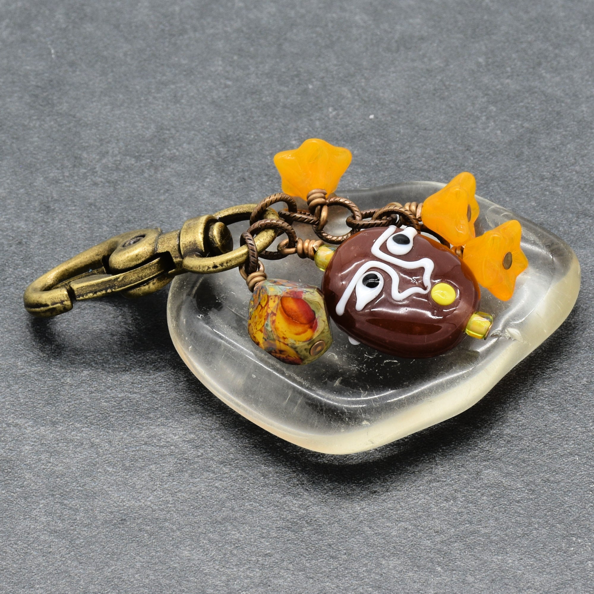 A handmade glass keychain featuring a clear, irregularly shaped glass base with a brown and white painted face, three small orange flowers, a patterned bead, and an antique bronze clasp, shown on a dark gray textured background - made with love in Colorado by Ruby Acorn.