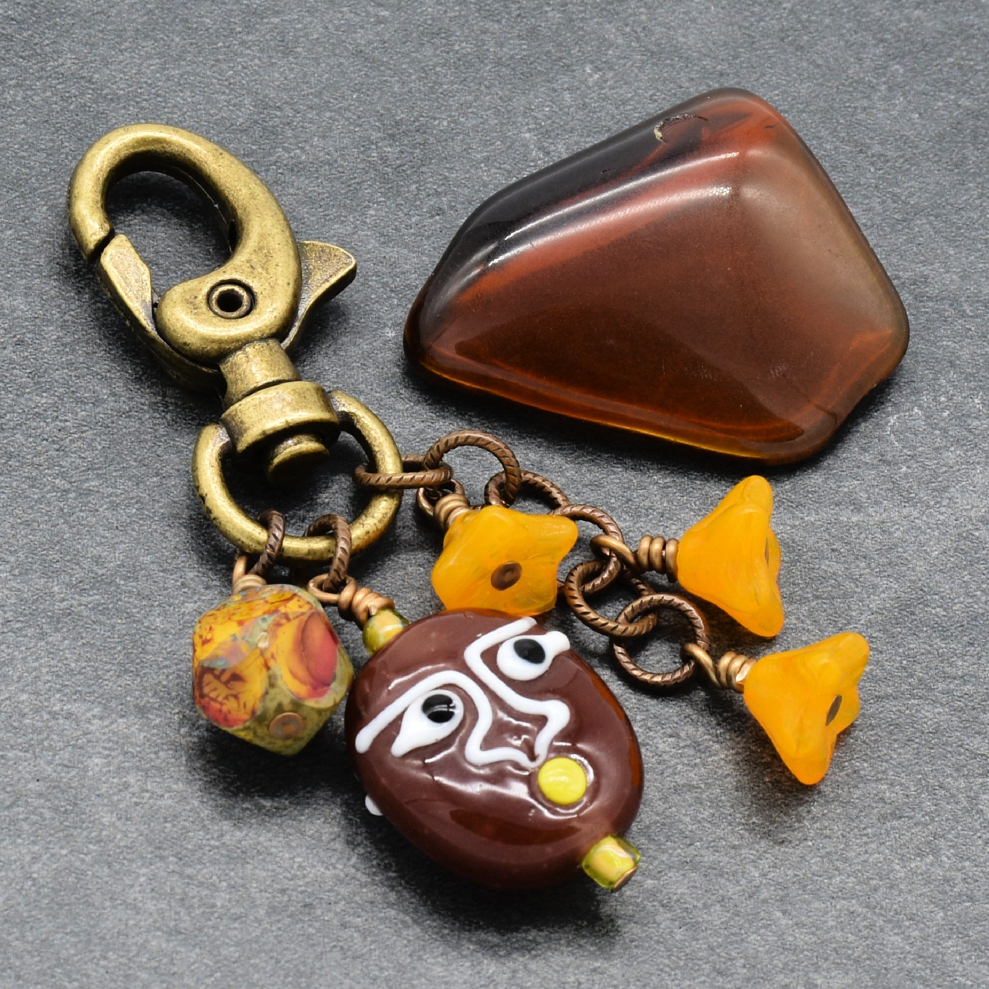 A handmade keychain charm featuring a brass clasp, a brown glass bead with white painted eyes, two yellow glass bell beads, and a marbled bead, all attached to a chain next to a polished brown glass stone on a gray textured background - Colorado made by Ruby Acorn.