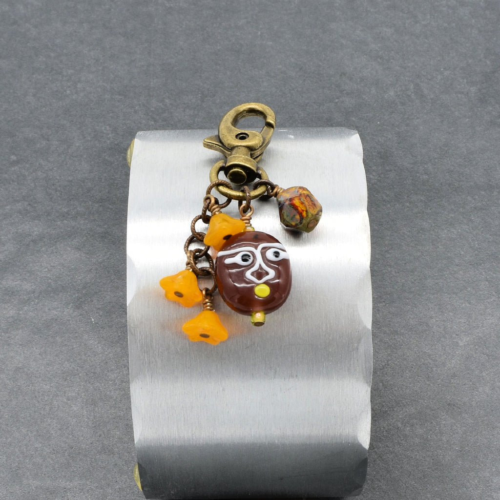 A brass-toned keychain or bag charm with a lobster clasp, dangling beads, and a brown oval pendant featuring a stylized face, displayed on a brushed metal stand against a gray background - made with love in Colorado by Ruby Acorn.