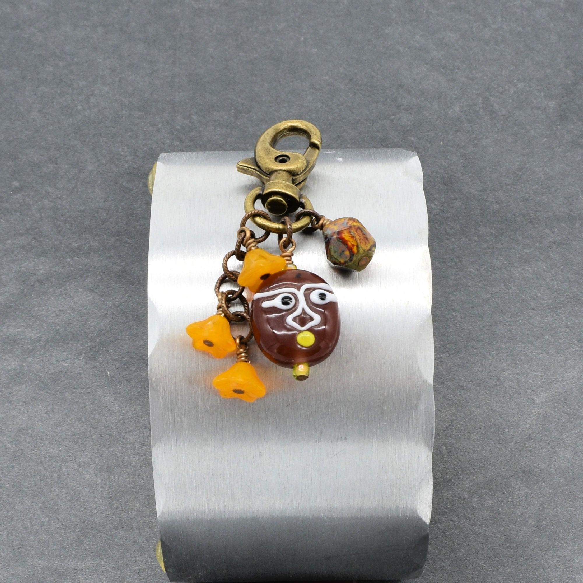 A brass-toned keychain or bag charm with a lobster clasp, dangling beads, and a brown oval pendant featuring a stylized face, displayed on a brushed metal stand against a gray background - made with love in Colorado by Ruby Acorn.