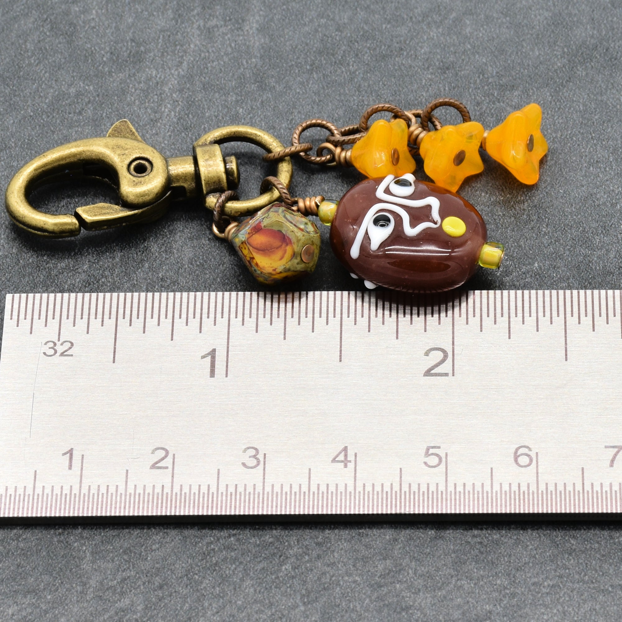 A handmade keychain or bag charm featuring a bronze-finished lobster clasp, a brown glass bead with white painted details, a small marbled bead, and three yellow flower-shaped beads, shown next to a metal ruler for scale on a gray background - made with love in Colorado by Ruby Acorn.