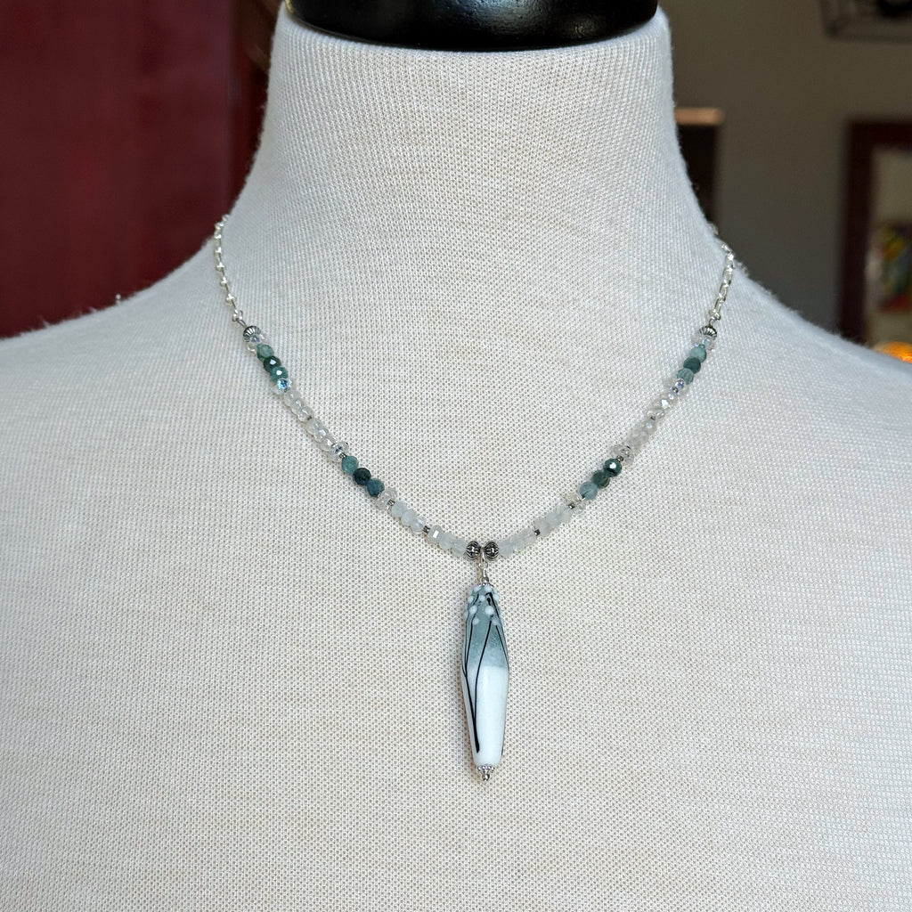 A beaded necklace with a long tapered barrel artisan lampwork glass pendant displayed on a cream fabric mannequin bust. The necklace features a silver-tone chain with clear faceted beads, small dark green beads, and the focal pendant.