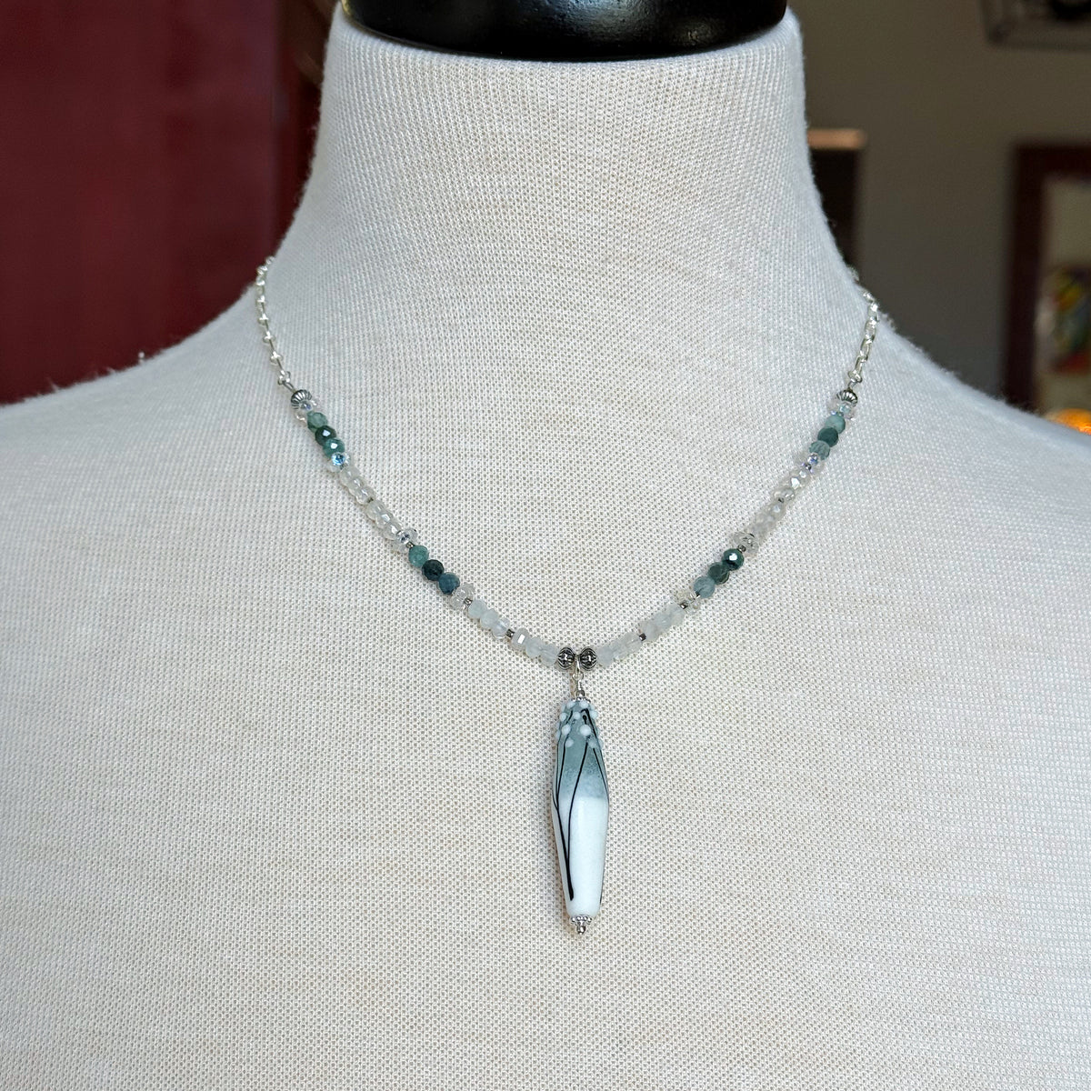 A beaded necklace with a long tapered barrel artisan lampwork glass pendant displayed on a cream fabric mannequin bust. The necklace features a silver-tone chain with clear faceted beads, small dark green beads, and the focal pendant.
