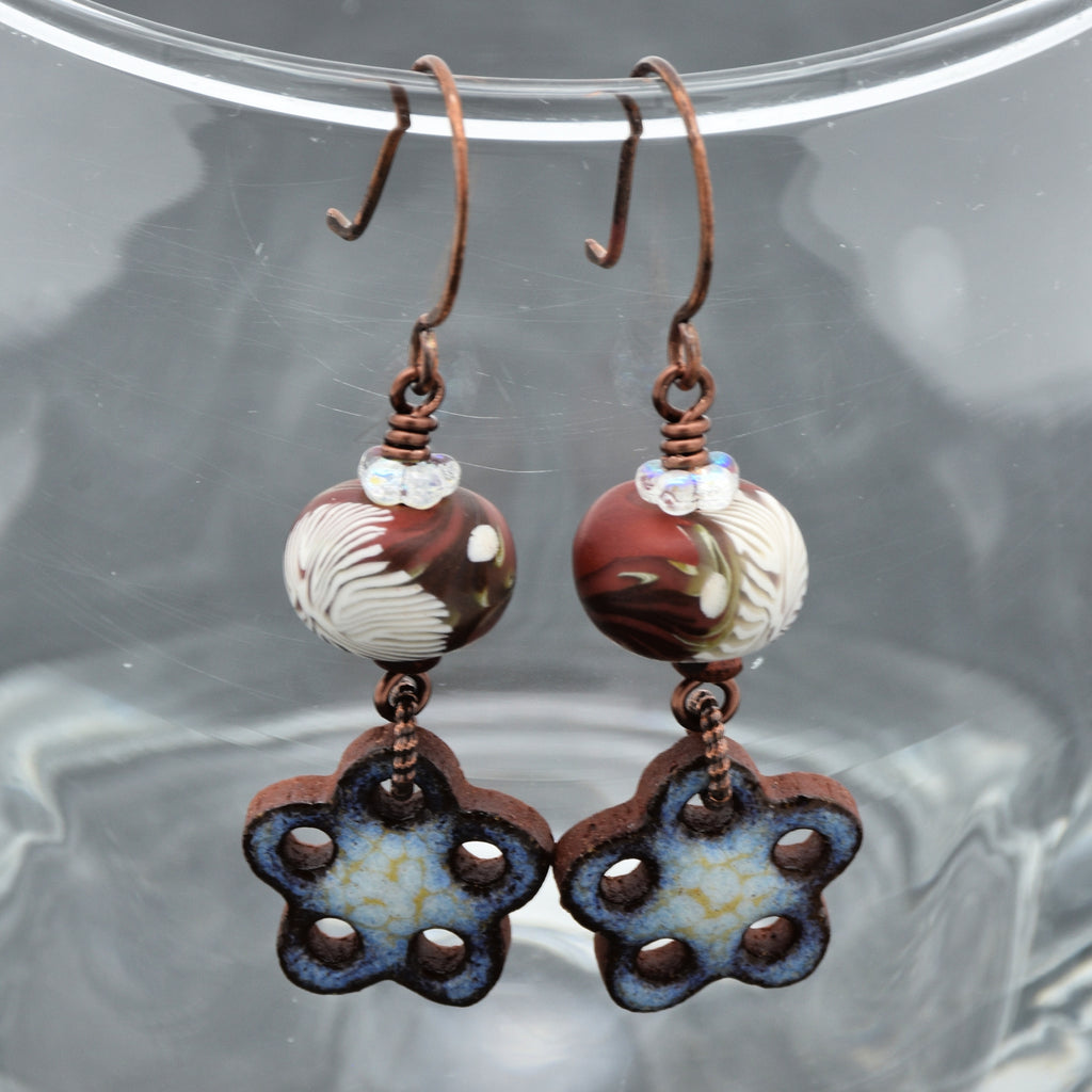 Pair of earrings with marbled beads and flower-shaped pendants on a reflective surface.