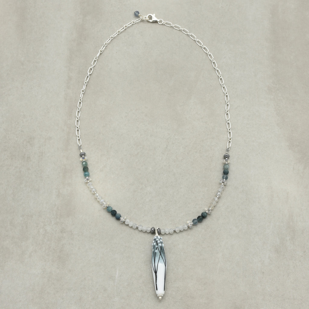 A delicate silver chain necklace featuring a central white teardrop pendant with blue-green branch detailing, flanked by small dark blue beads and clear crystal spacers, displayed on a neutral beige background.