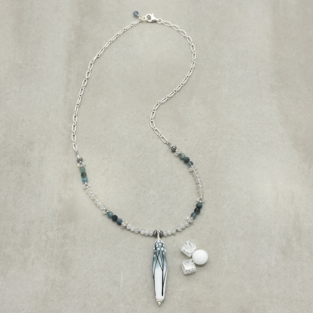 A sterling silver beaded necklace with a long barrel artisan lampwork glass pendant featuring blue and white branch patterns, displayed on a textured beige background. The necklace includes small dark blue-gray beads, and clear faceted beads.
