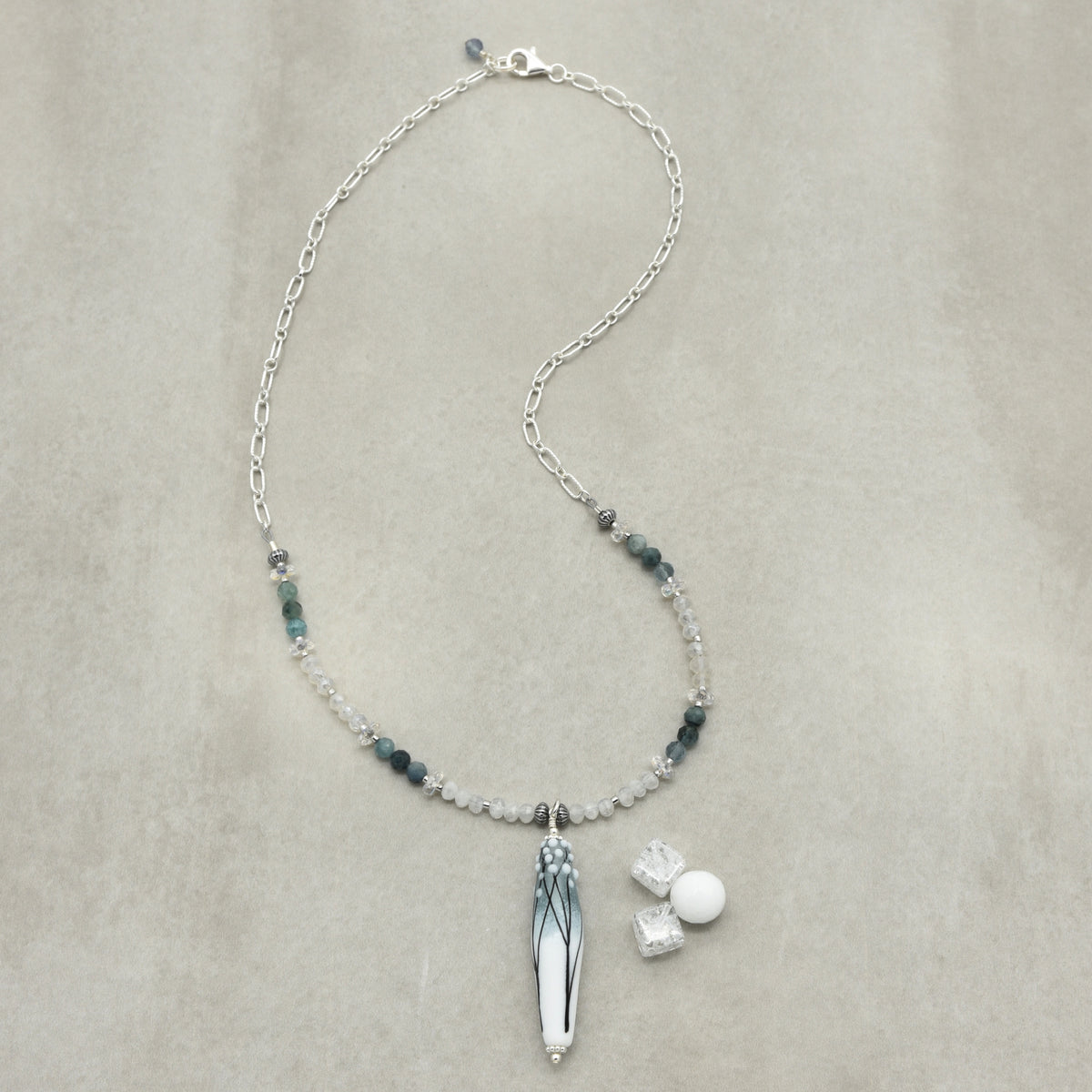 A sterling silver beaded necklace with a long barrel artisan lampwork glass pendant featuring blue and white branch patterns, displayed on a textured beige background. The necklace includes small dark blue-gray beads, and clear faceted beads.
