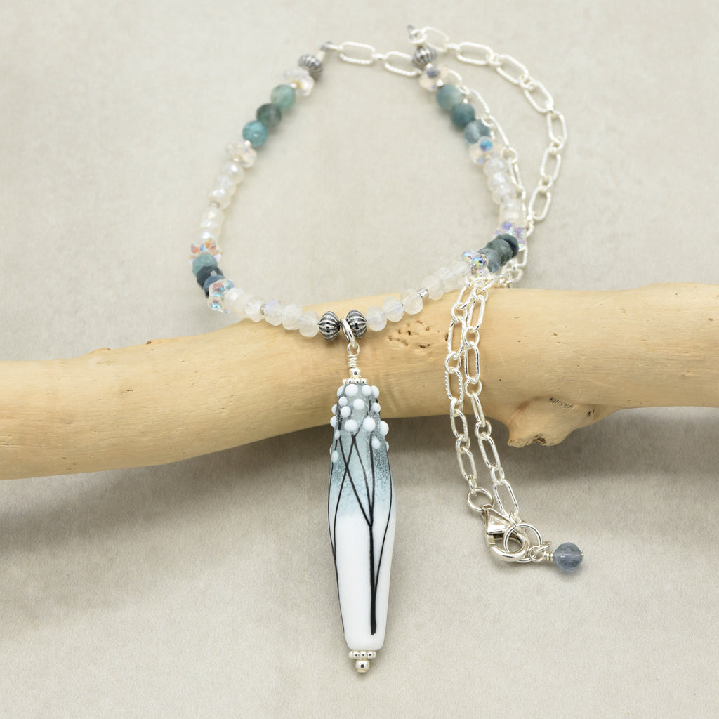 A beaded necklace with a long barrel artisan lampwork glass pendant featuring a blue and white gradient with black branch details, displayed on a light wood branch against a neutral beige background.