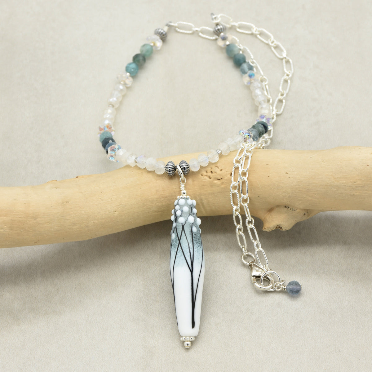 A beaded necklace with a long barrel artisan lampwork glass pendant featuring a blue and white gradient with black branch details, displayed on a light wood branch against a neutral beige background.