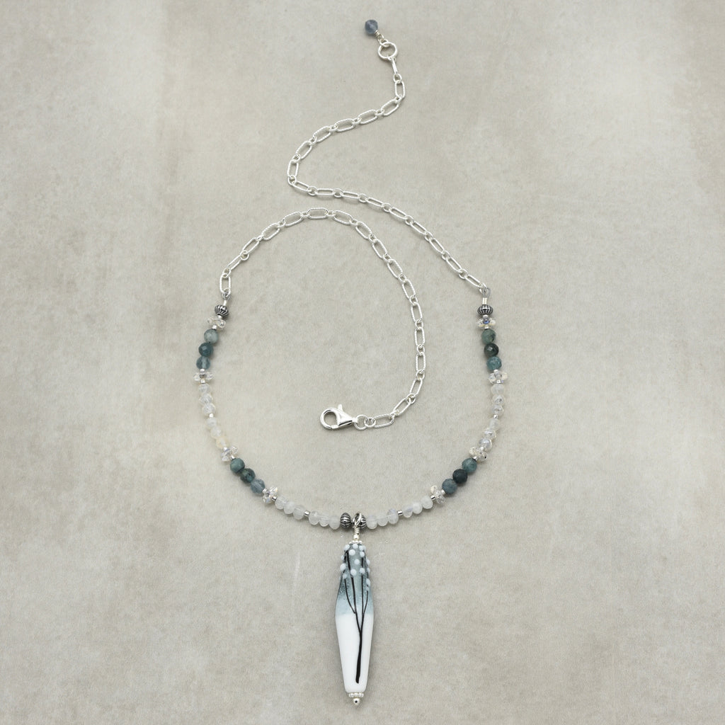 A delicate silver chain necklace featuring a central white thin tapered barrel artisan lampwork glass pendant with a black and gray leaf design, accented by small dark green and clear beads on a neutral beige background.