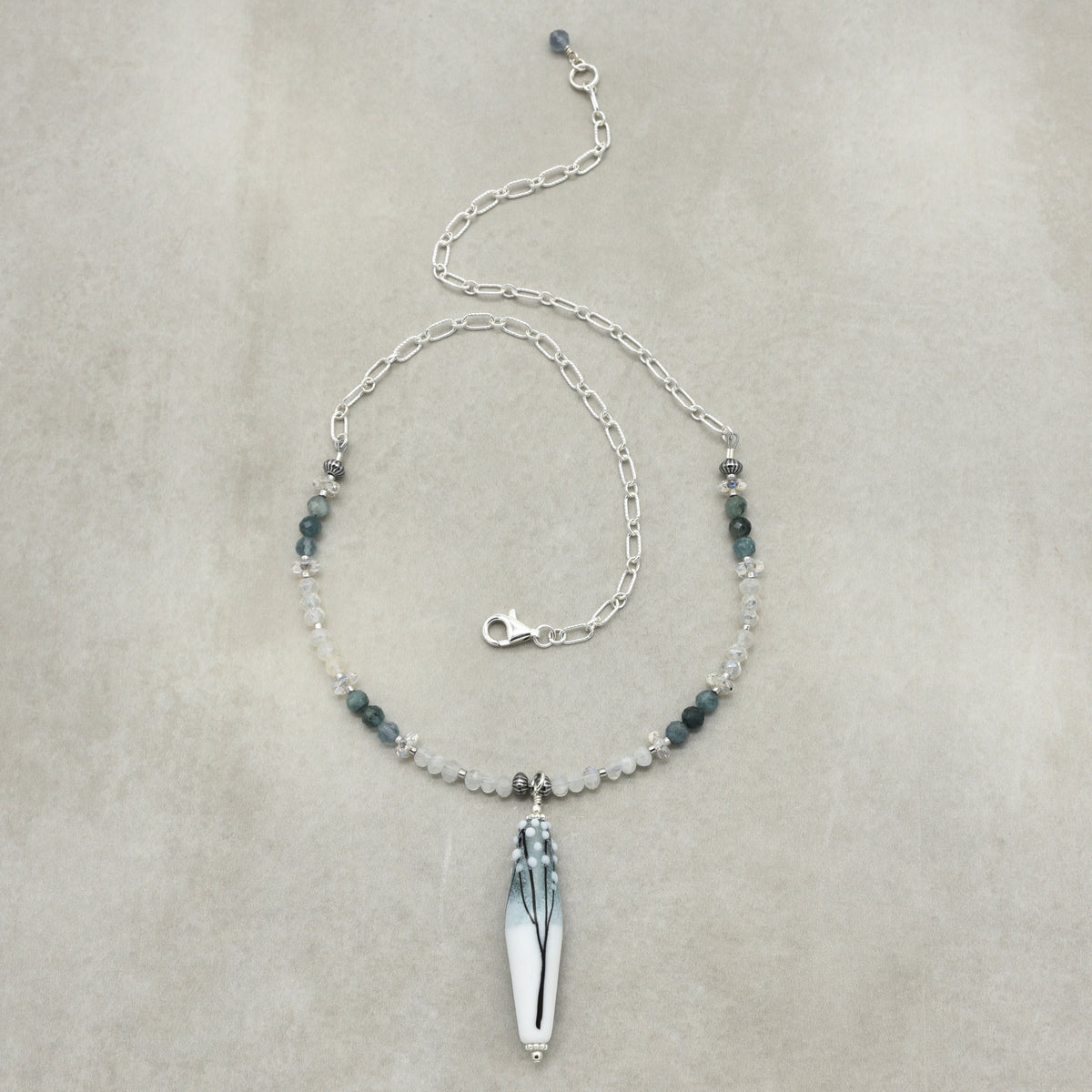 A delicate silver chain necklace featuring a central white thin tapered barrel artisan lampwork glass pendant with a black and gray leaf design, accented by small dark green and clear beads on a neutral beige background.