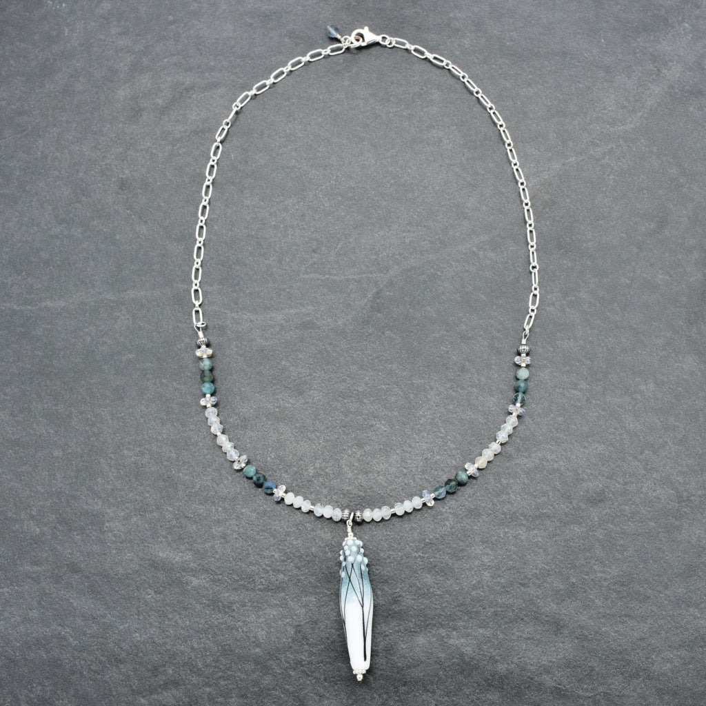 A silver chain necklace featuring a central  pendant with blue and white marbled detailing, flanked by small white and blue beads on a dark gray textured background.