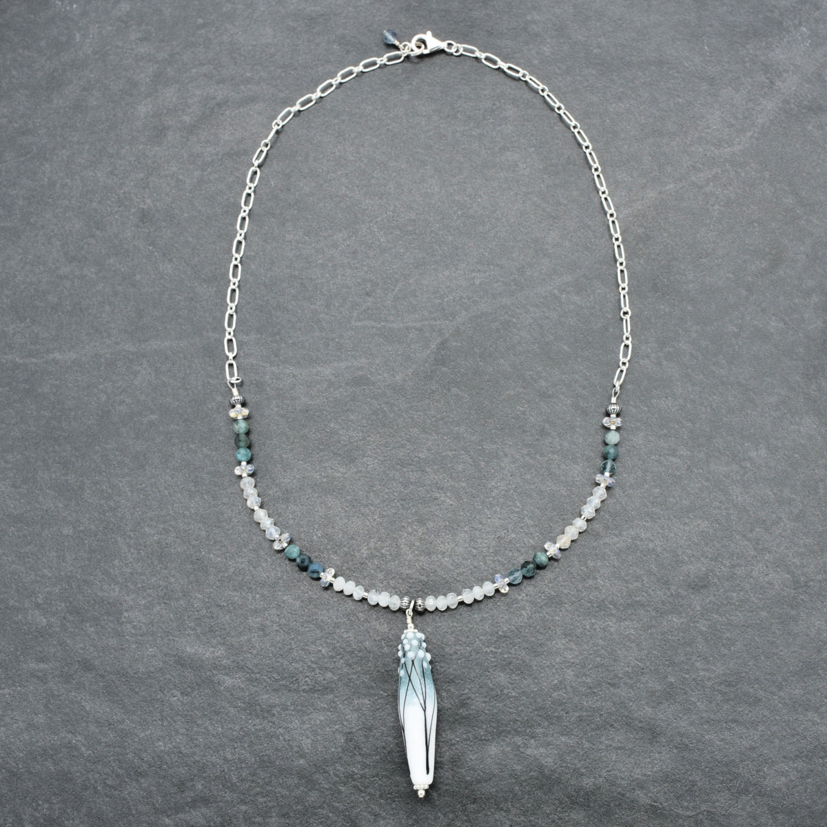 A silver chain necklace featuring a central  pendant with blue and white marbled detailing, flanked by small white and blue beads on a dark gray textured background.