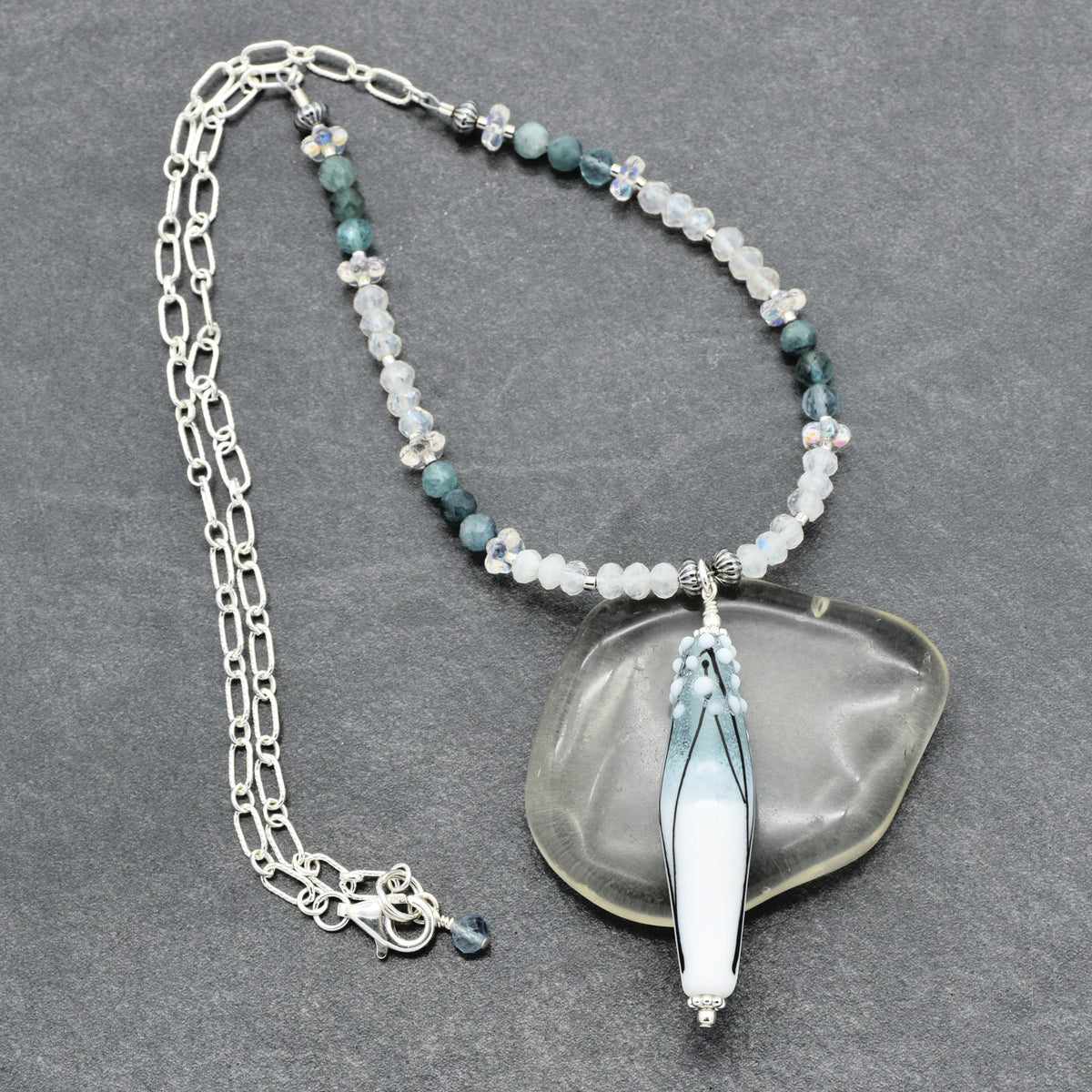 A beaded necklace with a large thin barrel shaped artisan lampwork glass pendant, displayed on a dark gray textured background. The necklace features a sterling silver paperclip chain, and white and light blue beads.