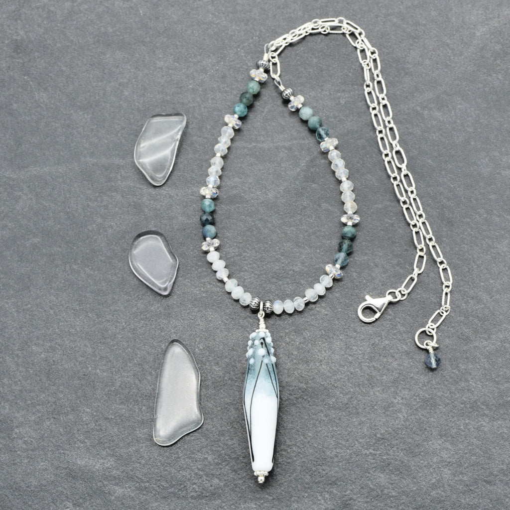 A beaded necklace with a long tapered barrel artisan lampwork glass pendant lies on a dark gray textured surface, alongside three clear, irregular-shaped stone pieces. The necklace features silver-toned chain and clasp, white and pale blue beads, and dark teal-green beads, with a central  pendant.