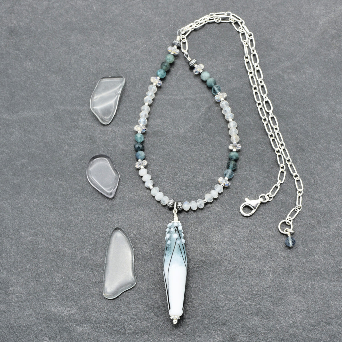 A beaded necklace with a long tapered barrel artisan lampwork glass pendant lies on a dark gray textured surface, alongside three clear, irregular-shaped stone pieces. The necklace features silver-toned chain and clasp, white and pale blue beads, and dark teal-green beads, with a central  pendant.