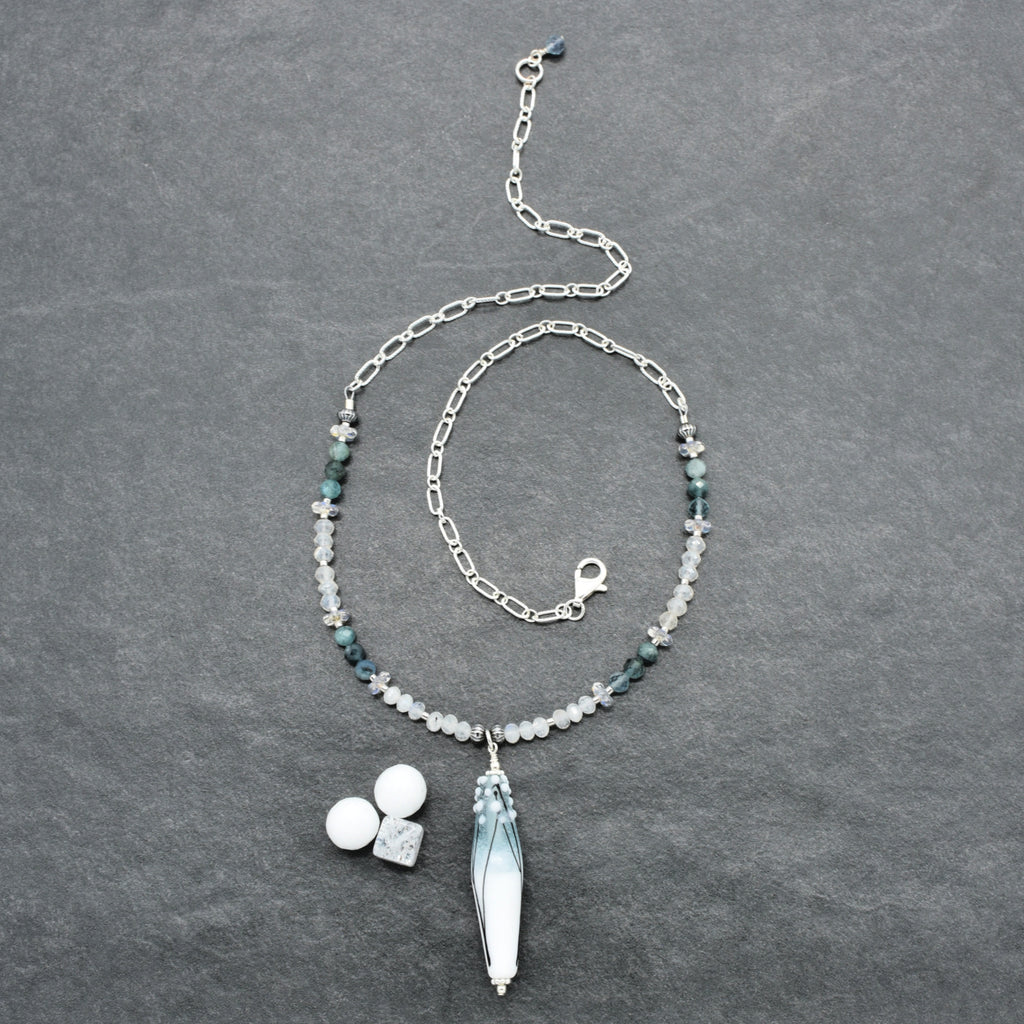 A silver-toned beaded necklace with a long barrel tapered artisan lampwork glass pendant, laid flat on a dark gray textured background. The necklace features small dark green and white beads, a silver link chain, a lobster clasp, and a large pendant with blue-green gradient detailing.