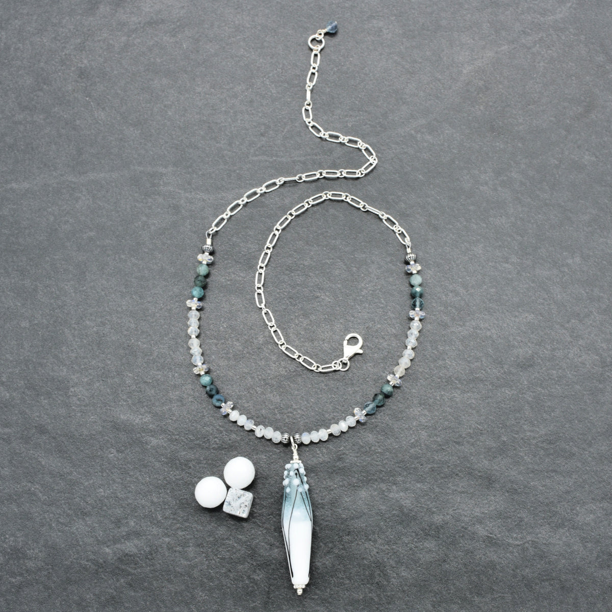 A silver-toned beaded necklace with a long barrel tapered artisan lampwork glass pendant, laid flat on a dark gray textured background. The necklace features small dark green and white beads, a silver link chain, a lobster clasp, and a large pendant with blue-green gradient detailing.