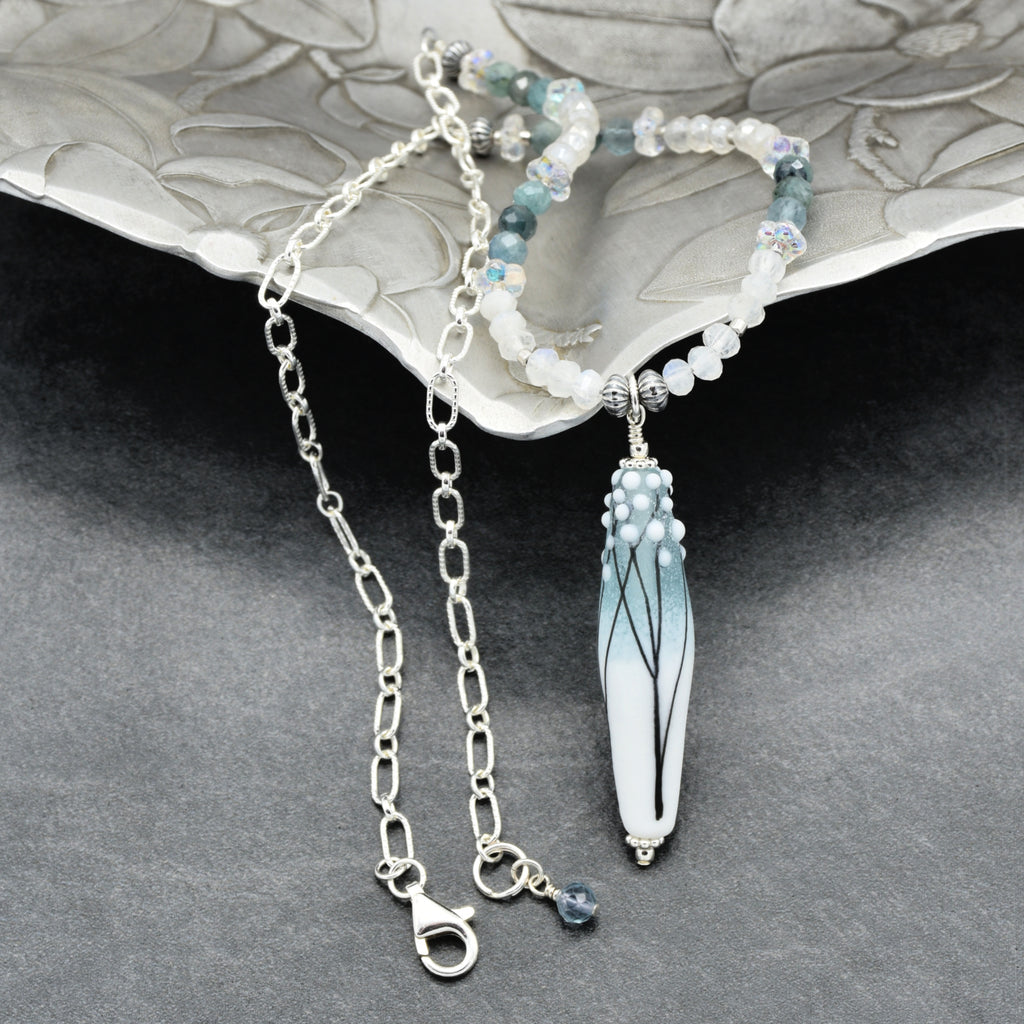 A beaded necklace with a long white and light blue glass pendant featuring black branches, displayed on a textured silver tray over a dark gray surface.