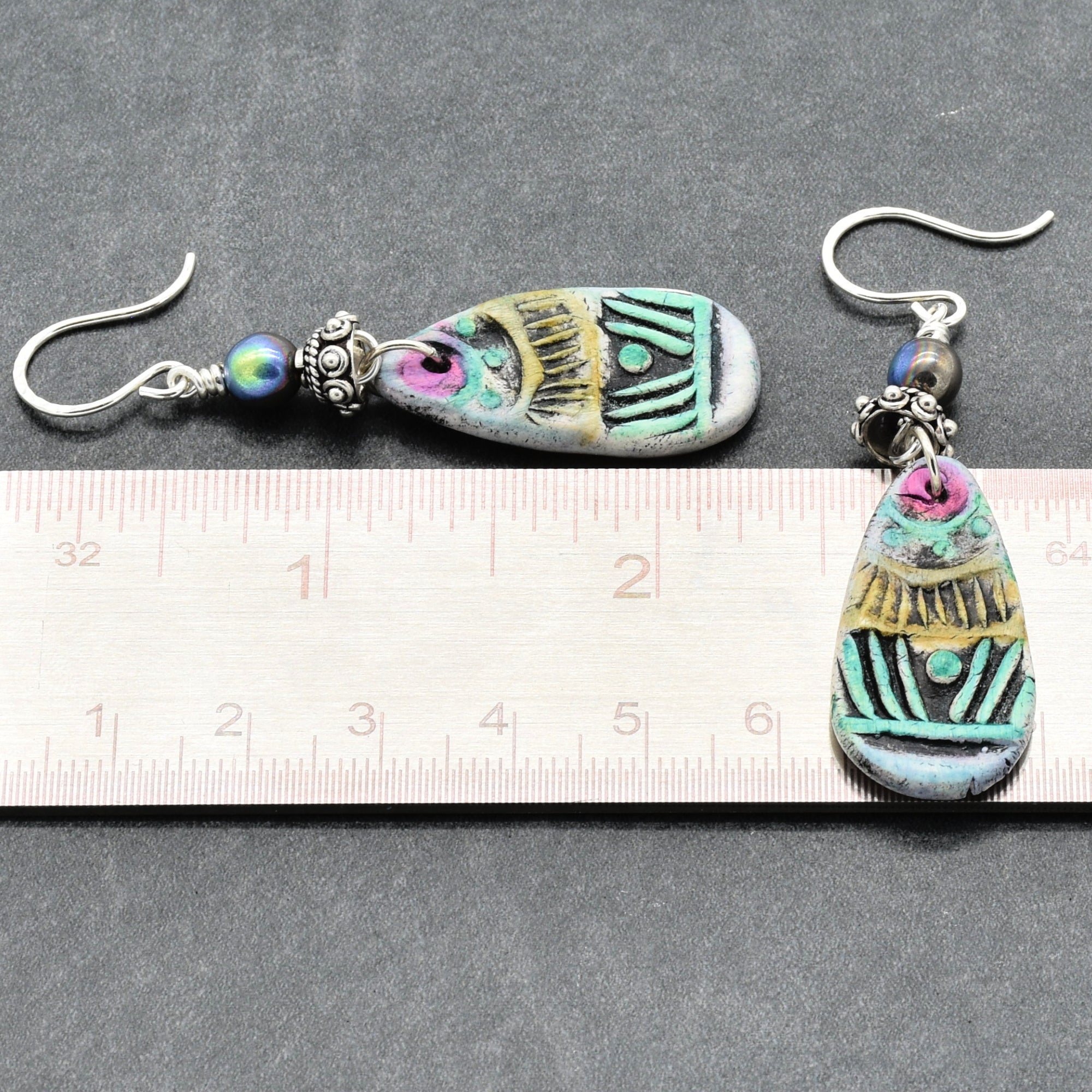 Pair of earrings with decorative elements on a ruler for scale, on a gray background