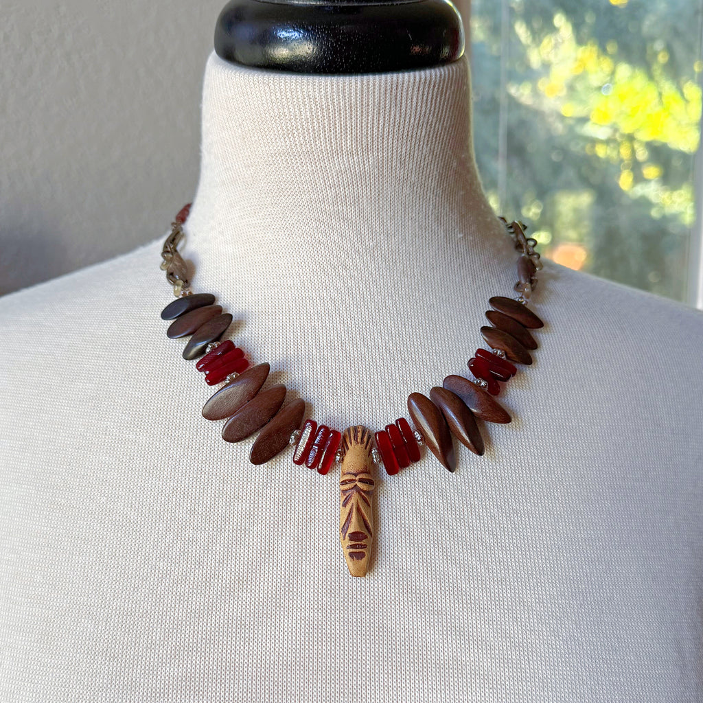 The image shows a necklace. It is made of brown, red, and beige beads, with a central beige pendant that has a face-like design carved into it. The necklace is displayed on a mannequin.