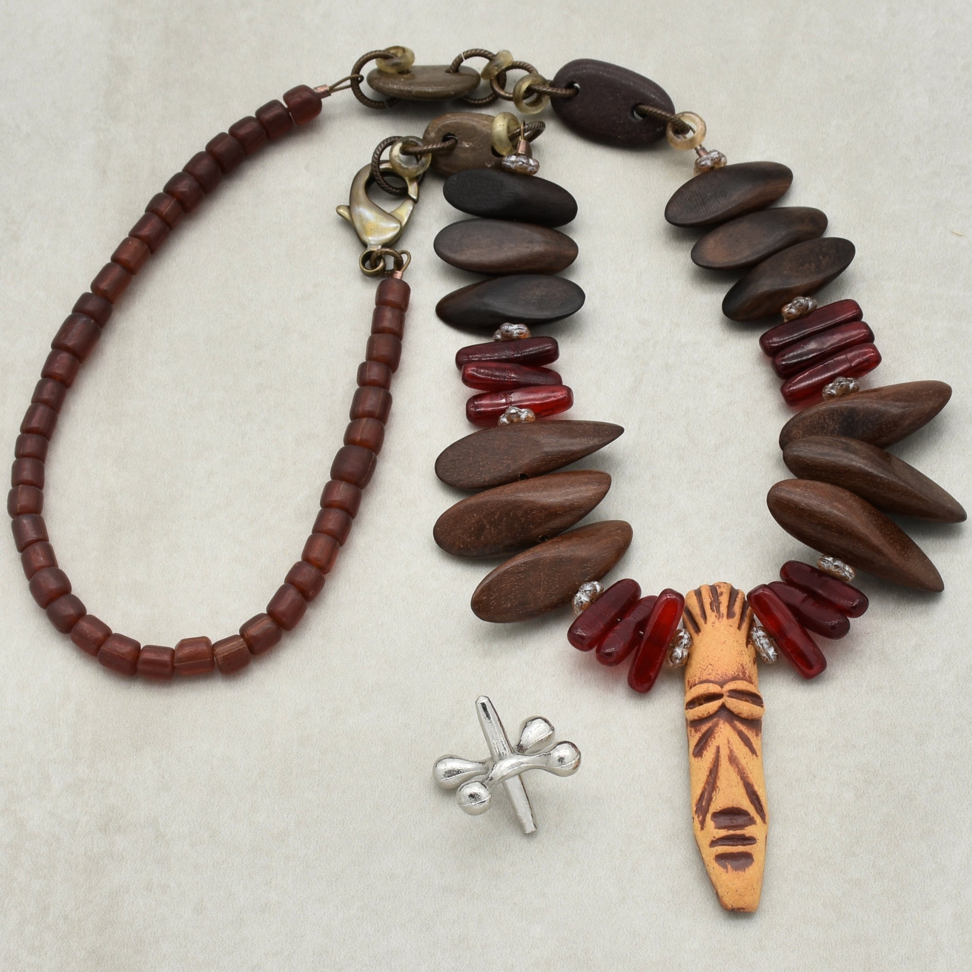 The image shows a necklace. It features a variety of beads, including reddish-brown cylindrical beads, dark wooden bead pieces, and red translucent beads. It also has a carved central pendant with a face-like design. There is a clasp visible at one end, confirming it as a necklace.
