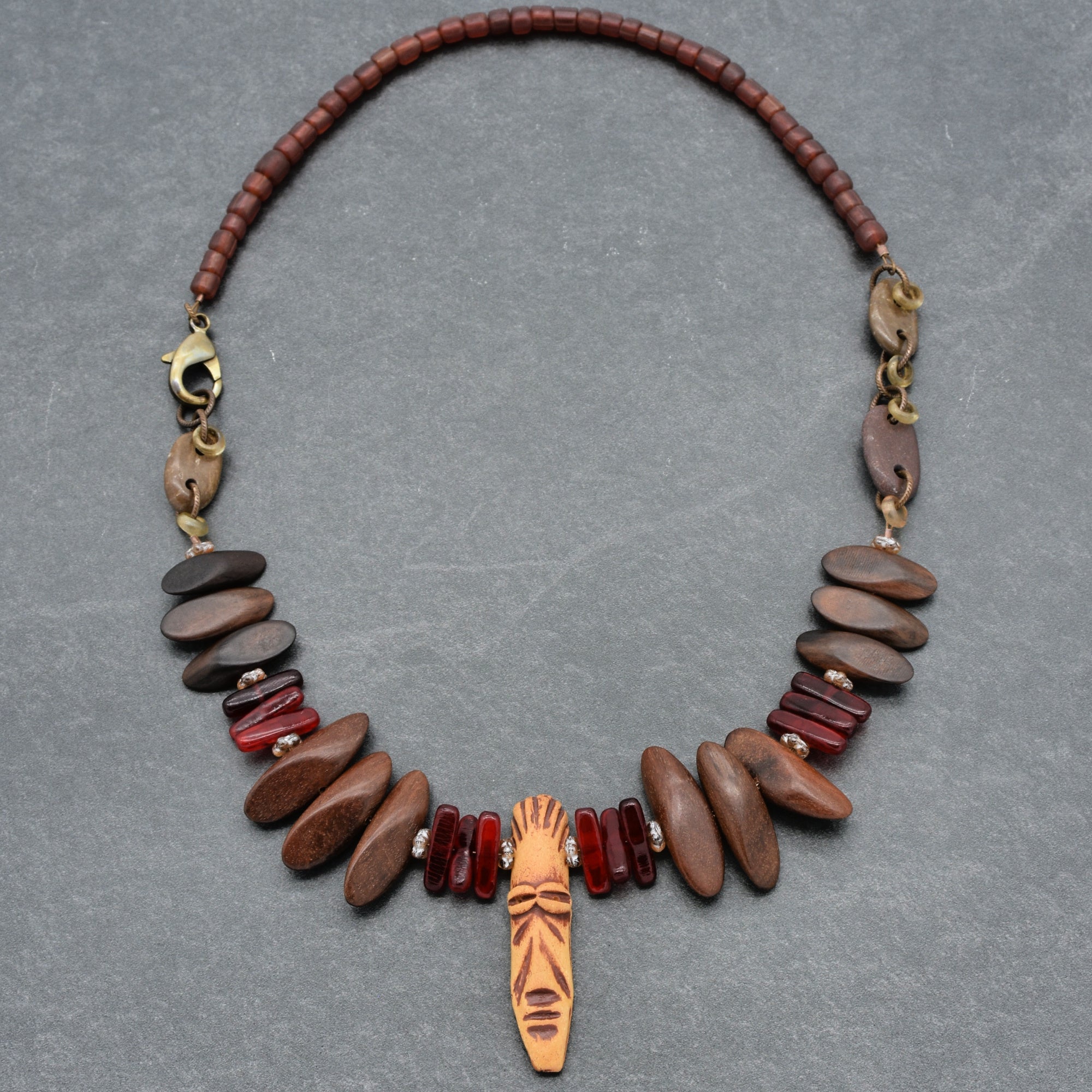 This image shows a necklace. It features a combination of brown, wooden beads, red beads, and a central carved wooden pendant with a face design. The necklace also has a metal clasp for fastening.