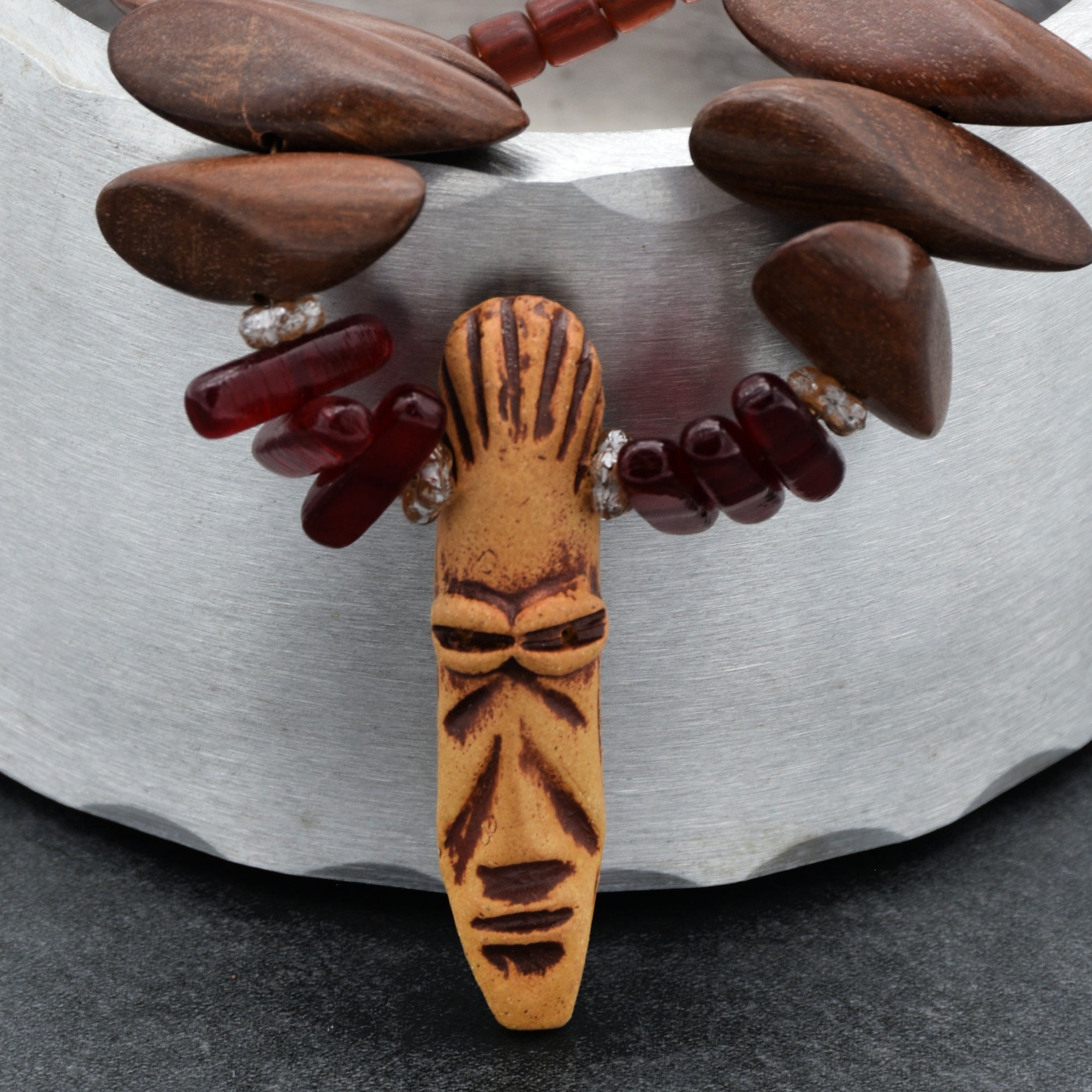 This image shows a necklace. The necklace features a carved face pendant, along with other beads made of wood and a reddish material. The shape and size of the pendant, along with the beads strung together, indicate that this is a necklace.