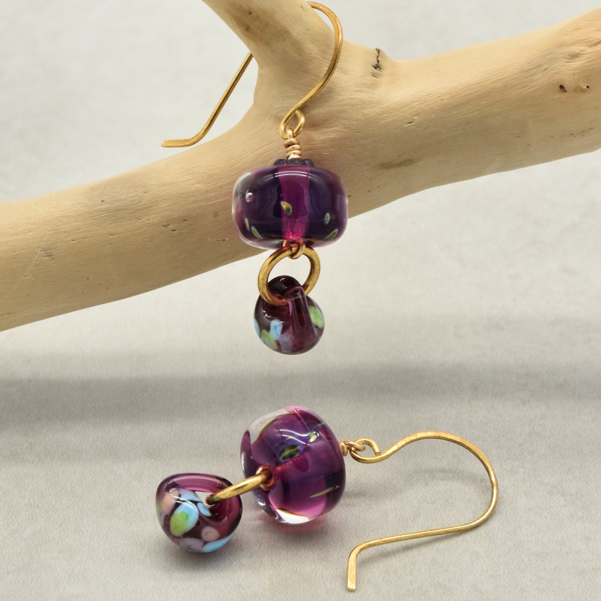 Purple glass bead earrings with brass hooks on a wooden branch and gray surface.