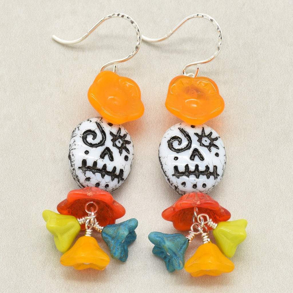 A pair of handmade sugar skull dangle earrings on a light beige textured background. The earrings feature silver fishhook wires, an orange translucent flower bead, a white round bead with black hand-drawn skull patterns, and a dangling cluster of red, yellow, blue, and orange flower beads - made with love in Colorado by Ruby Acorn.