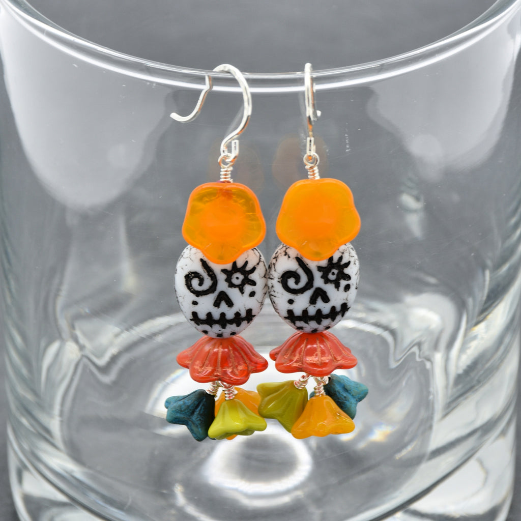 A pair of colorful sugar skull dangle earrings hanging in front of a clear glass tumbler. The earrings feature orange flower beads, white beads with black skull patterns, red flower charms, and small green and yellow flower charms at the bottom, suspended from silver fishhook wires - made with love in Colorado by Ruby Acorn.