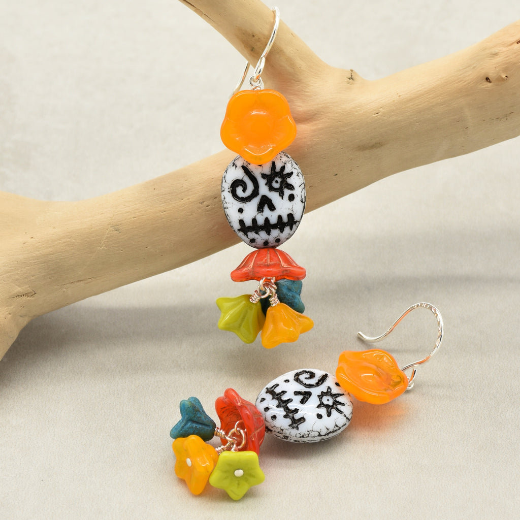 A pair of dangling earrings displayed on a light wood branch against a neutral beige background. The earrings feature a white stone bead with black skull and crossbones details, colorful flower charms in orange, yellow, green, and blue, and a small orange flower charm - made with love in Colorado by Ruby Acorn.