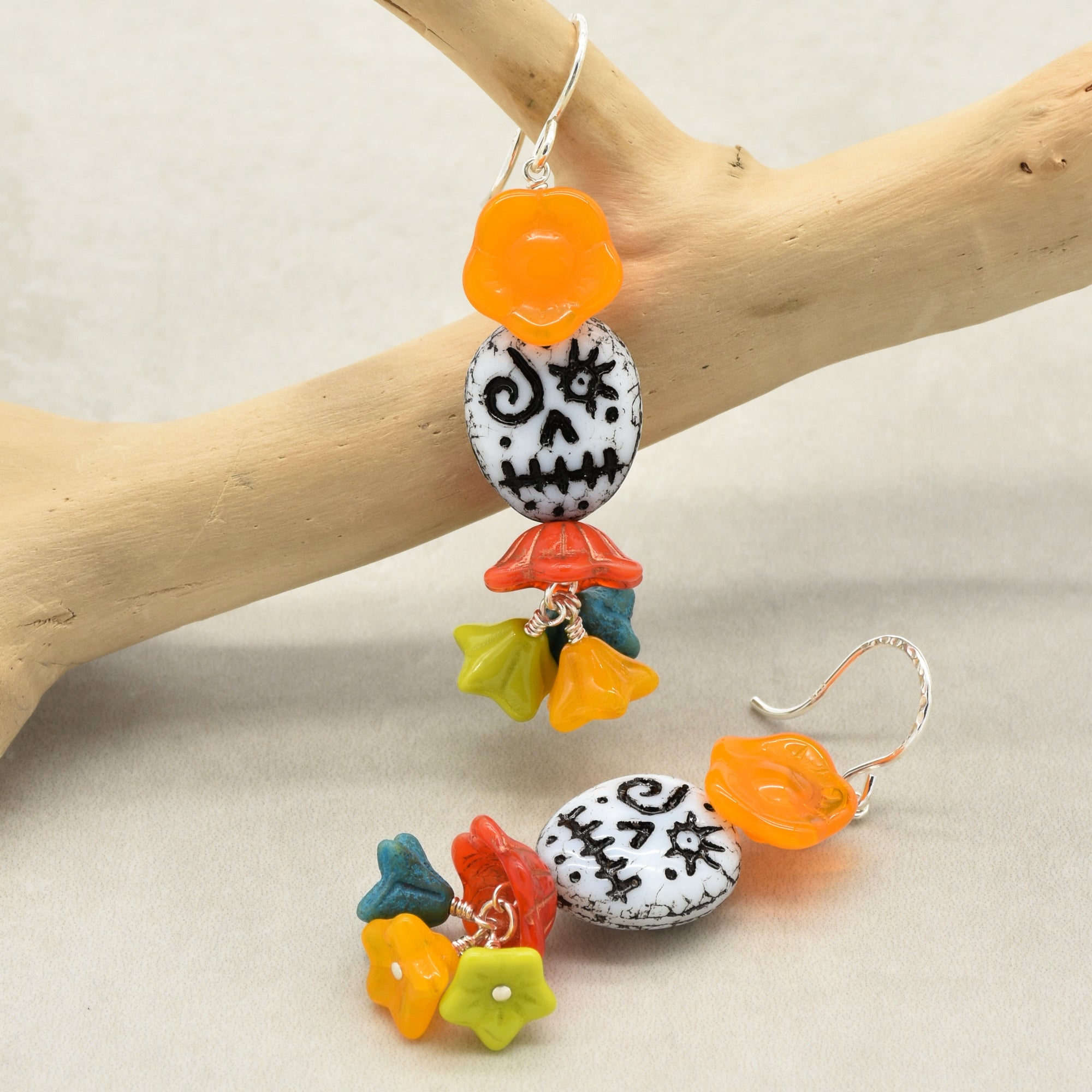 A pair of dangling earrings displayed on a light wood branch against a neutral beige background. The earrings feature a white stone bead with black skull and crossbones details, colorful flower charms in orange, yellow, green, and blue, and a small orange flower charm - made with love in Colorado by Ruby Acorn.