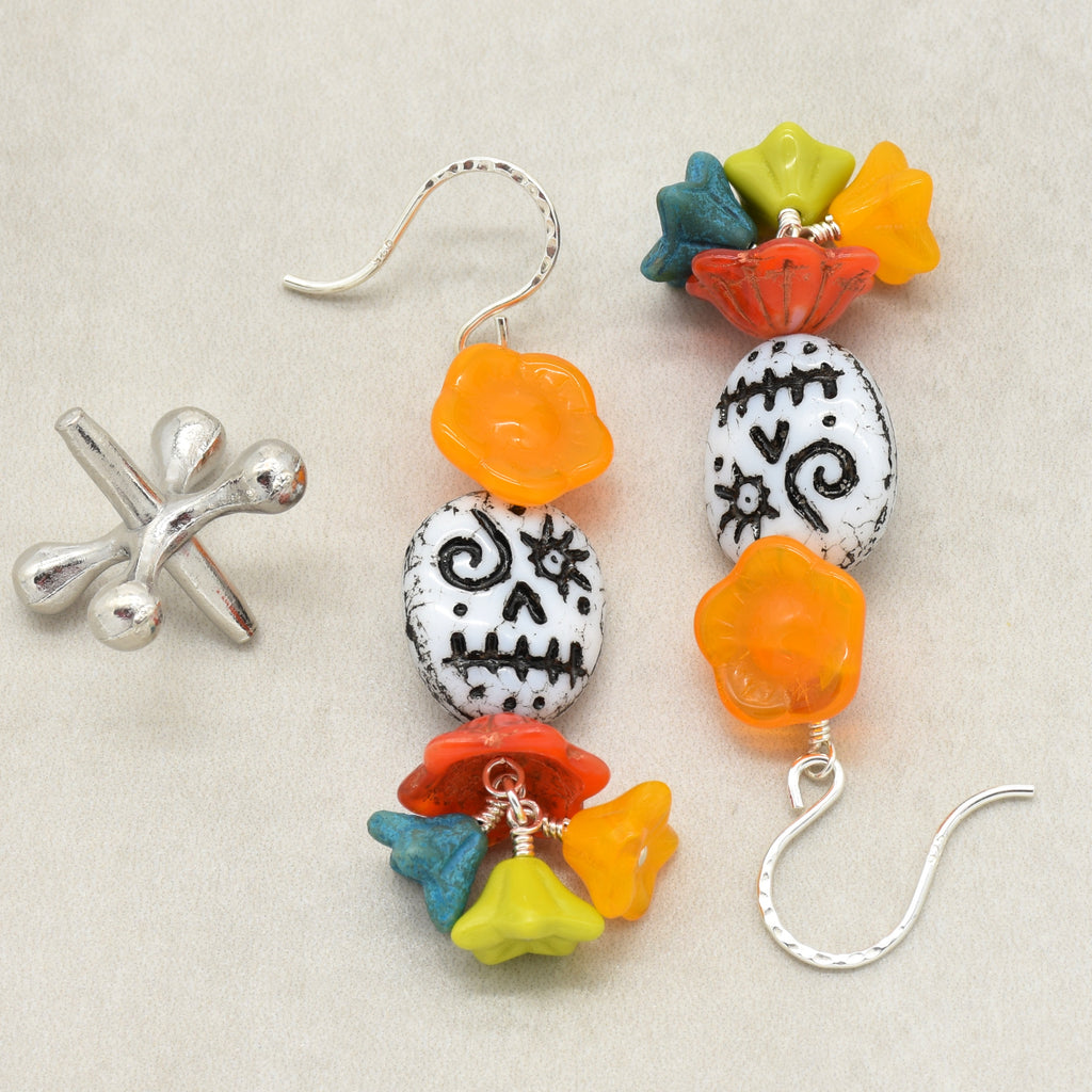 A pair of colorful beaded dangle earrings with silver fishhook wires, displayed on a neutral beige background. The earrings feature white sugar skull beads with black patterns, orange flower beads, and small multicolored star and flower charms in red, yellow, and blue - made with love in Colorado by Ruby Acorn.
