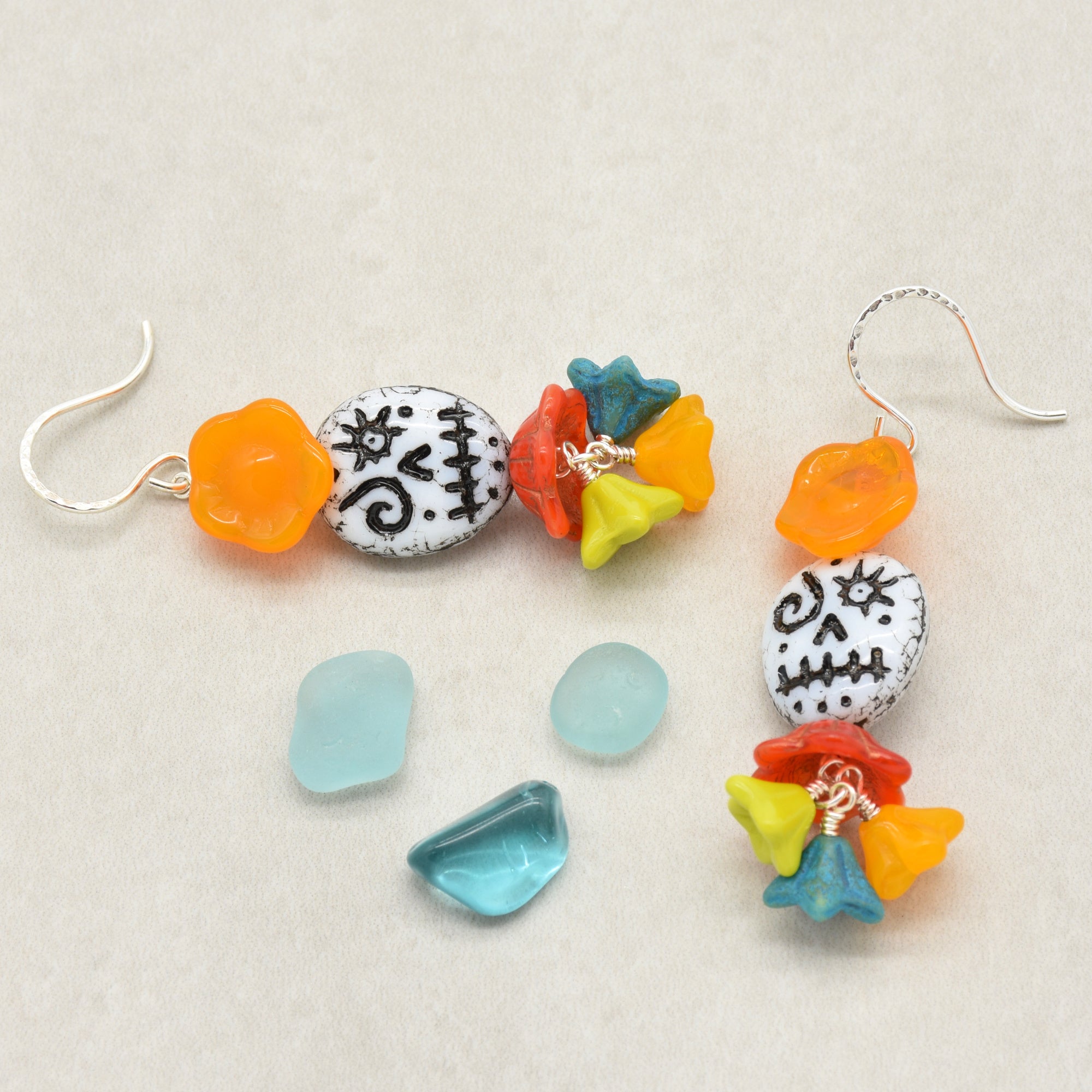 A pair of handmade beaded dangle earrings with colorful glass beads and silver fishhooks, displayed alongside loose matching beads on a neutral beige background - made with love in Colorado by Ruby Acorn.