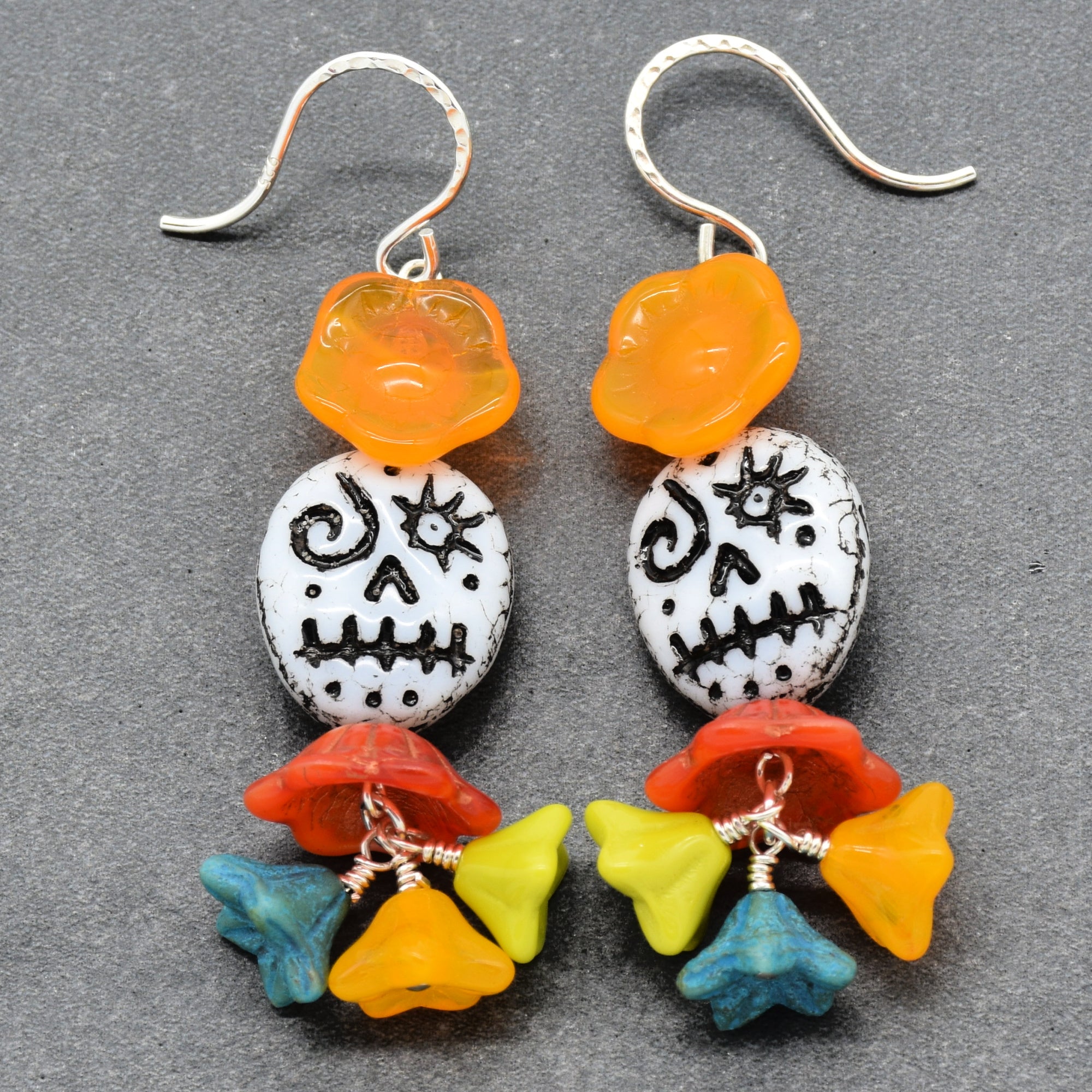 A pair of handmade dangle earrings featuring silver-tone fishhook wires, translucent orange flower beads, white beads with black sugar skull designs, and colorful flower charms in red, yellow, green, and blue at the bottom, displayed on a dark gray textured background - made with love in Colorado by Ruby Acorn.