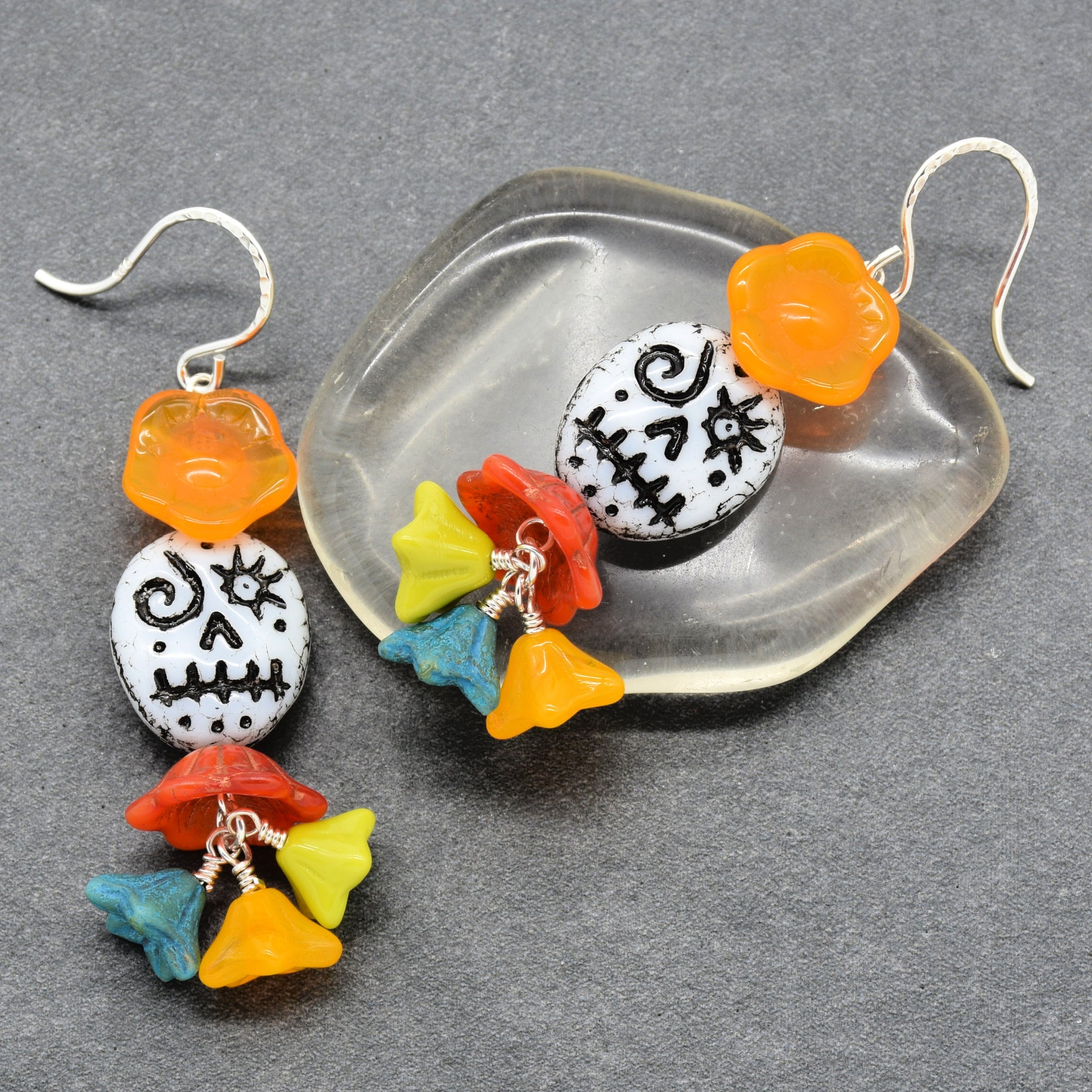 A pair of dangling earrings displayed on a gray surface, with one earring hanging flat and the other resting on a clear acrylic tray. The earrings feature a white sugar skull-style markings, an orange flower-shaped bead, and dangling multicolored bellflower charms in yellow, blue, red, and orange attached by silver wire - made with love in Colorado by Ruby Acorn.