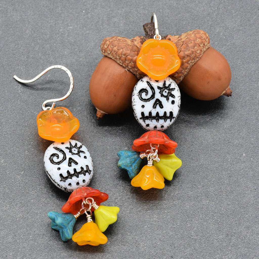 A pair of handmade dangle earrings with silver fishhook wires, featuring white beads with black skull designs, orange flower charms, and colorful dangling beads in red, yellow, blue, and green. The earrings are displayed against a dark gray background with decorative acorns - made with love in Colorado by Ruby Acorn.