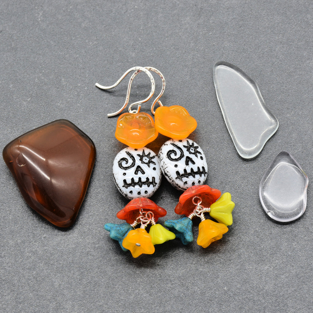 A pair of handmade dangle earrings displayed on a gray textured background, accompanied by two clear glass pieces and a polished brown stone. The earrings feature orange flower-shaped beads, white beads with black skull designs, red and orange flower charms, and yellow and blue charms - made with love in Colorado by Ruby Acorn.