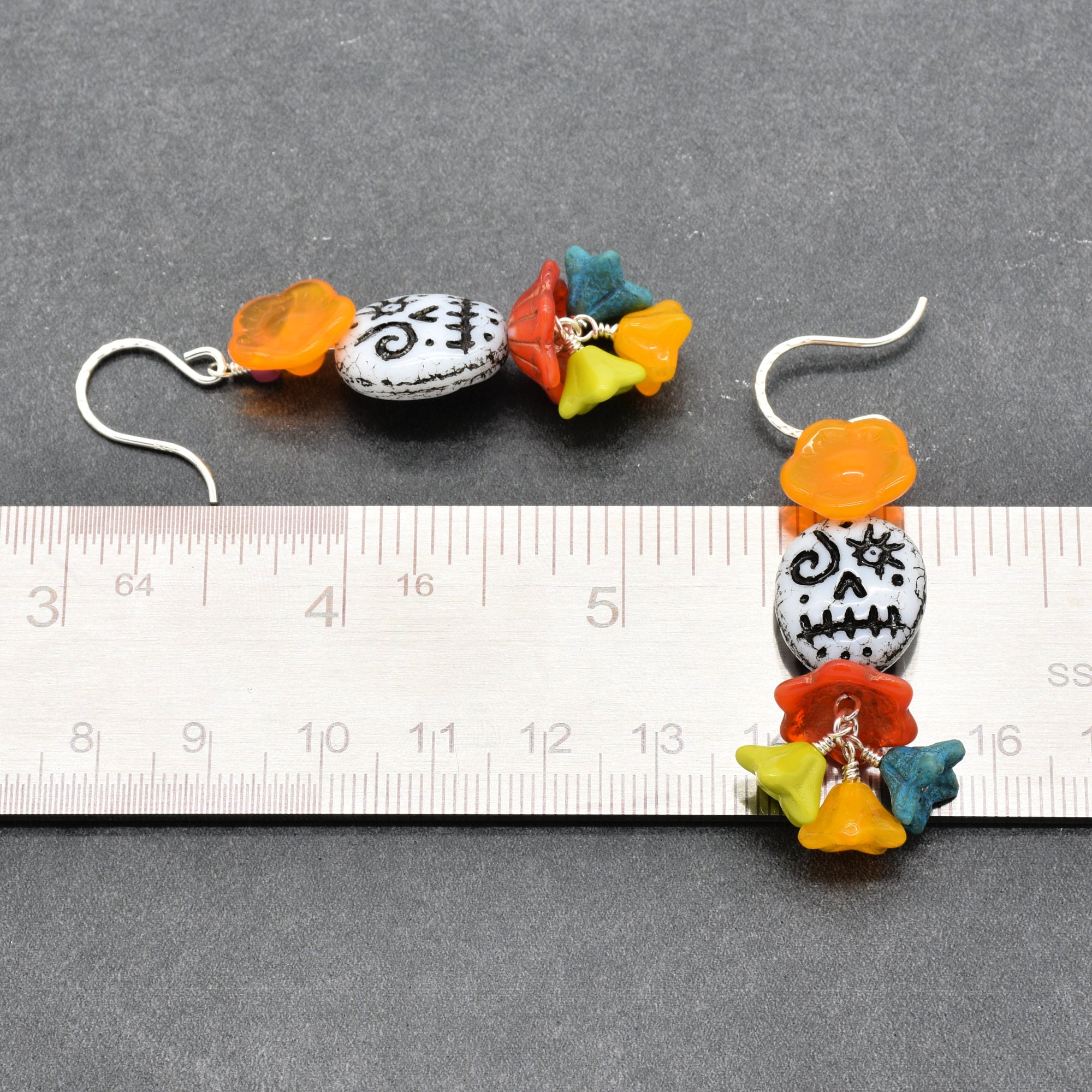 A pair of handmade beaded dangle earrings displayed on a dark gray textured background with a metal ruler for scale. The earrings feature a white round bead with black swirl patterns, colorful flower beads in orange, red, yellow, and teal, and silver-tone hooks - made with love in Colorado by Ruby Acorn.